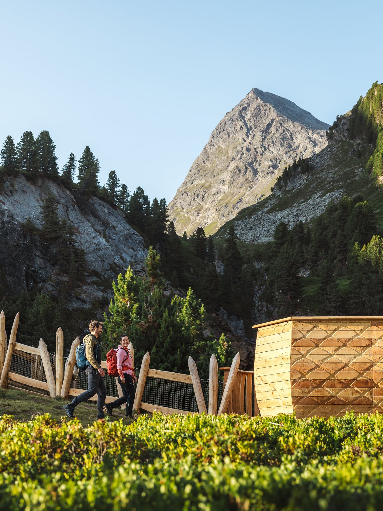 Two hikers navigate a nature trail surrounded by lush green mountains and wooden structures