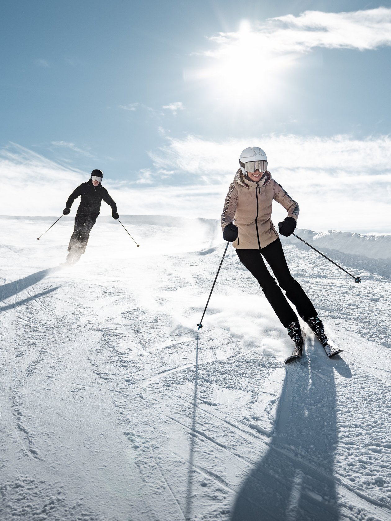Zwei Skifahrer fahren eine schneebedeckte Piste bei Sonnenschein unter blauem Himmel hinunter.