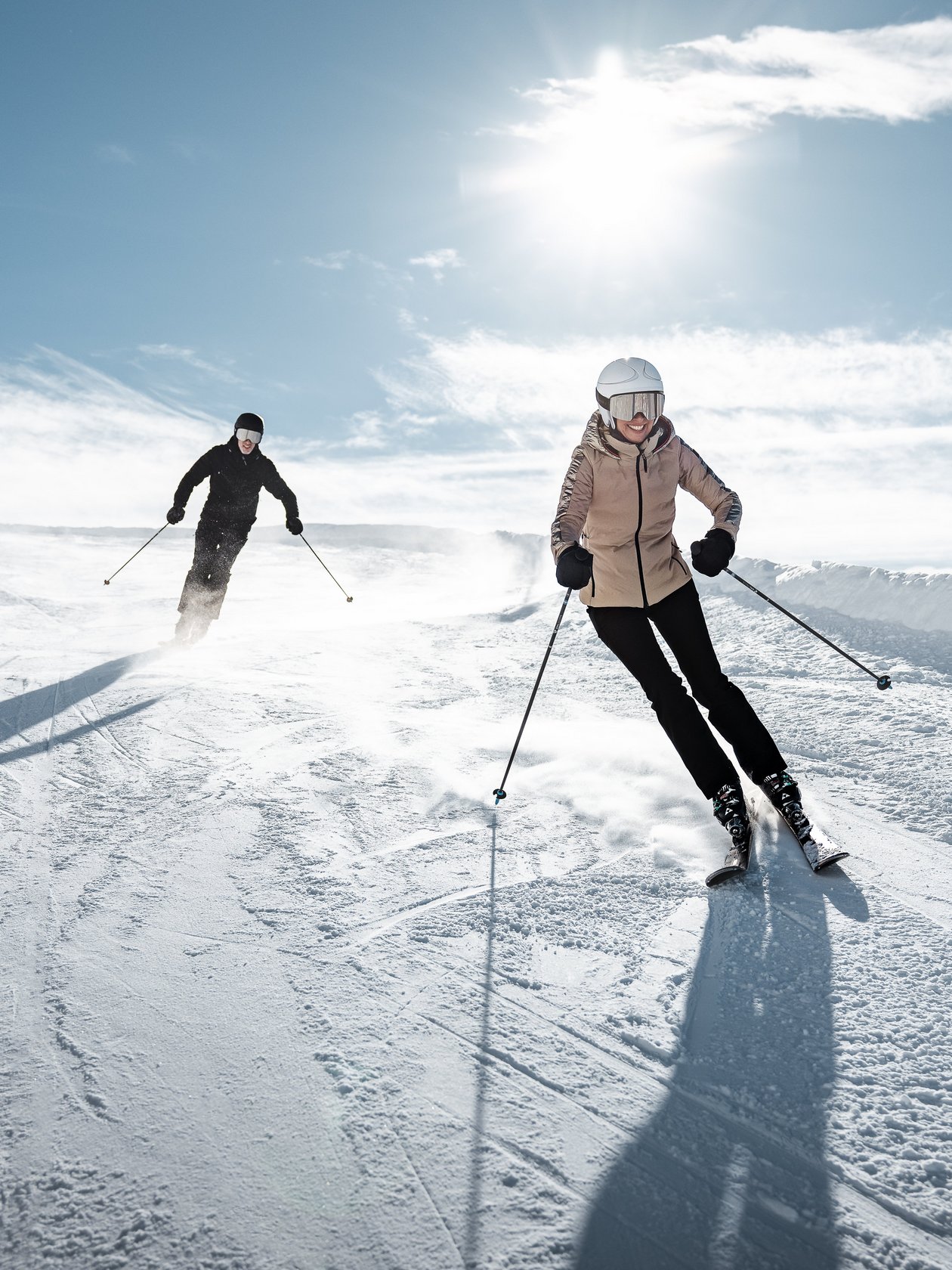 Zwei Skifahrer fahren eine schneebedeckte Piste bei Sonnenschein unter blauem Himmel hinunter.