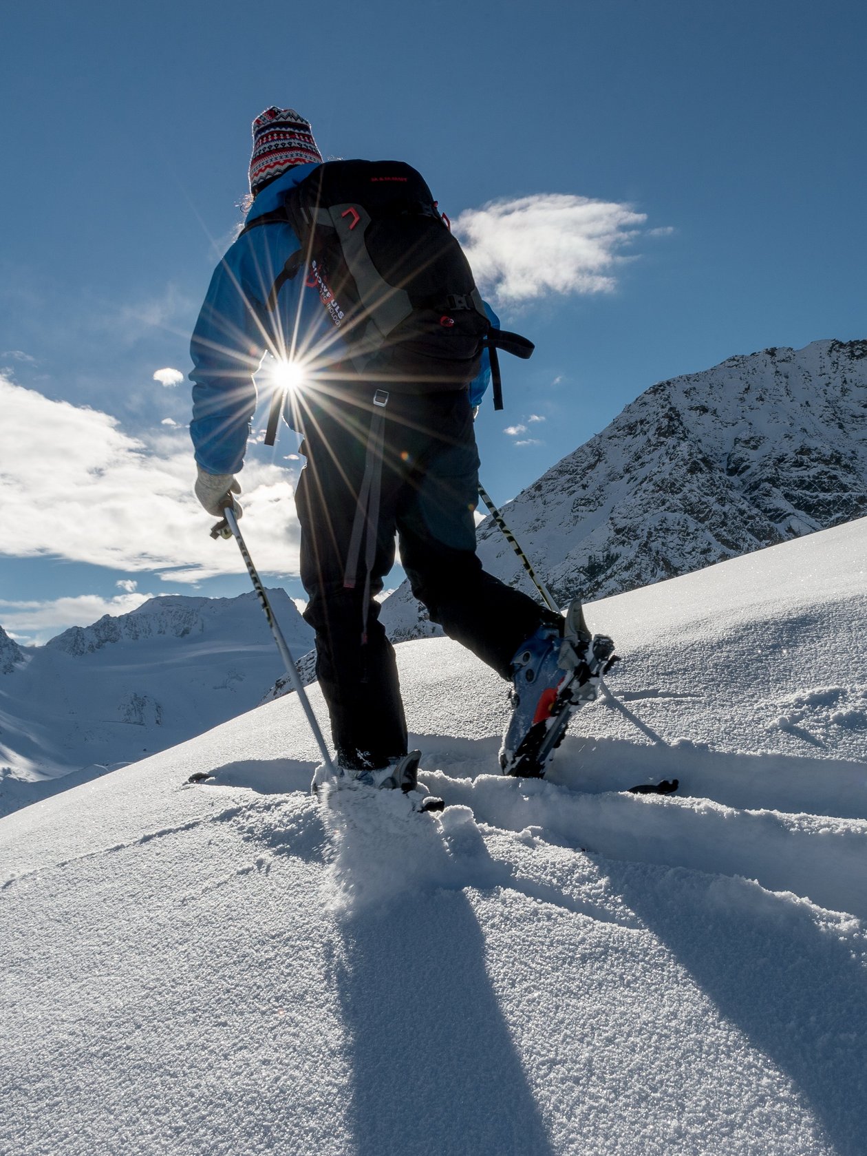 Eine Person erklimmt einen verschneiten Berg mit Sonnenlicht im Hintergrund, umgeben von schneebedeckter Landschaft.