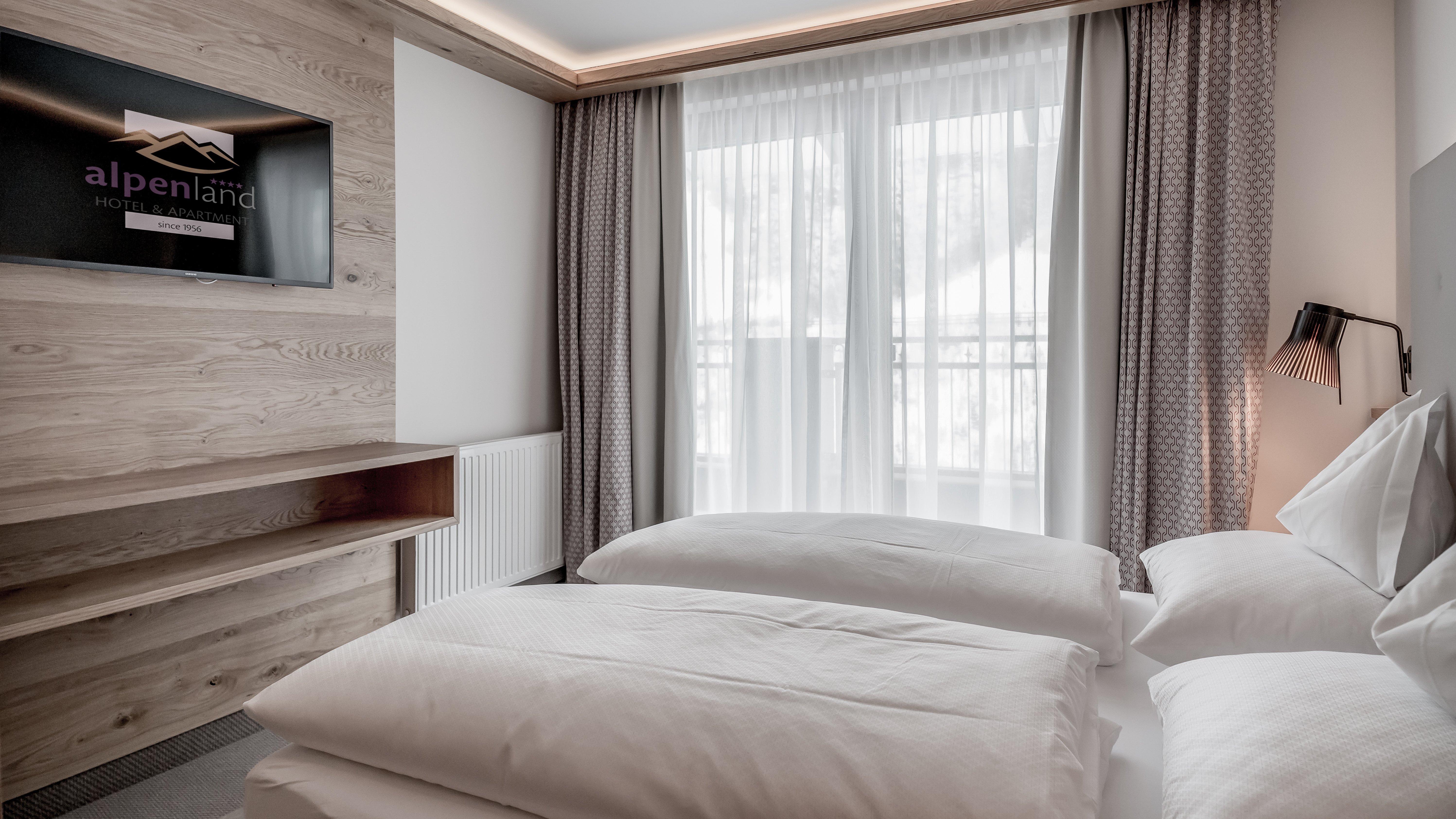 Modernes Hotelzimmer mit zwei Betten Hotelzimmer mit zwei Betten, Wandfernseher und großen Fenstern mit Vorhängen im Alpenland Hotel.
