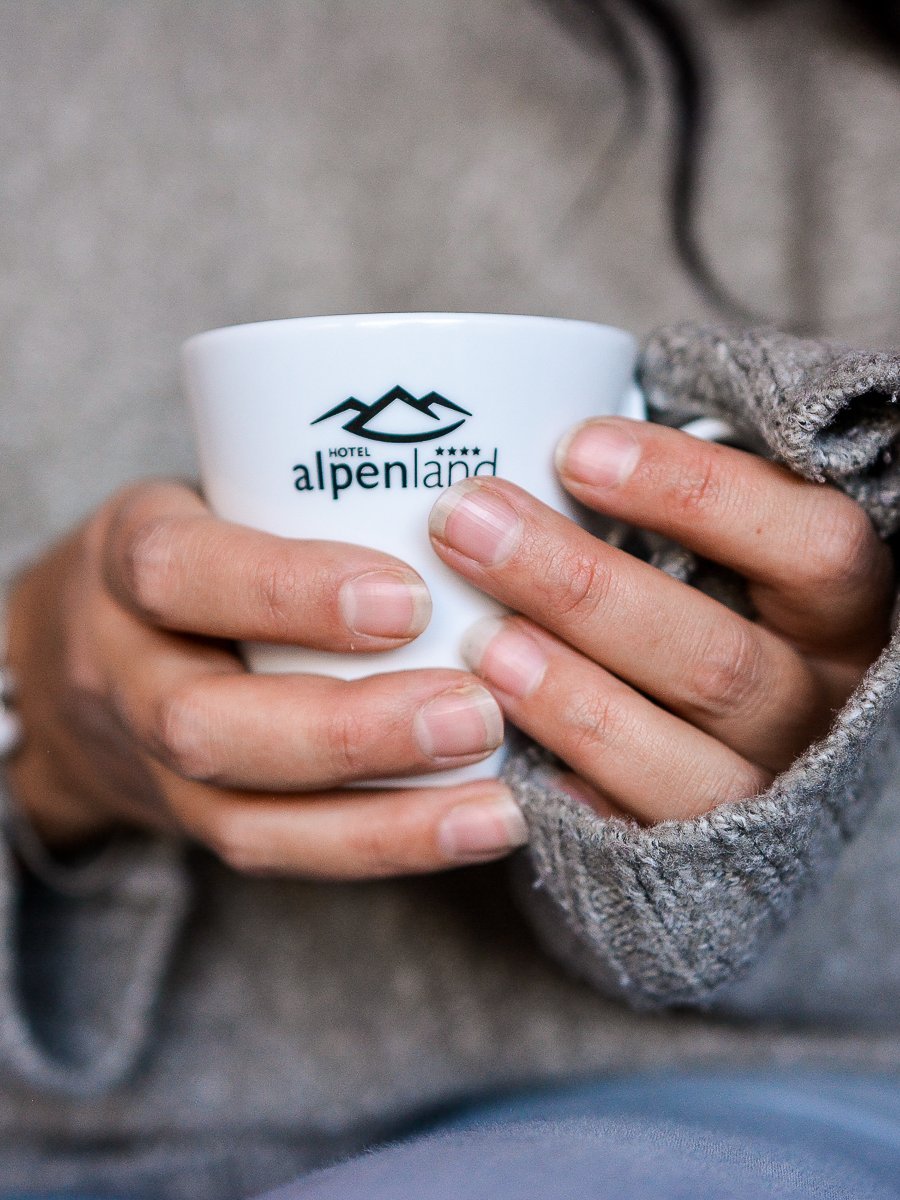 Kaffeegenuss im Alpenland Person hält eine weiße Kaffeetasse mit Hotel Alpenland Logo und trägt einen gemütlichen Pullover.
