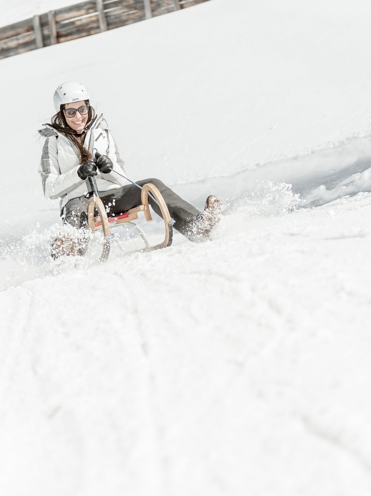 Two people sledding down a snowy slope with trees in the background and bright snow all around