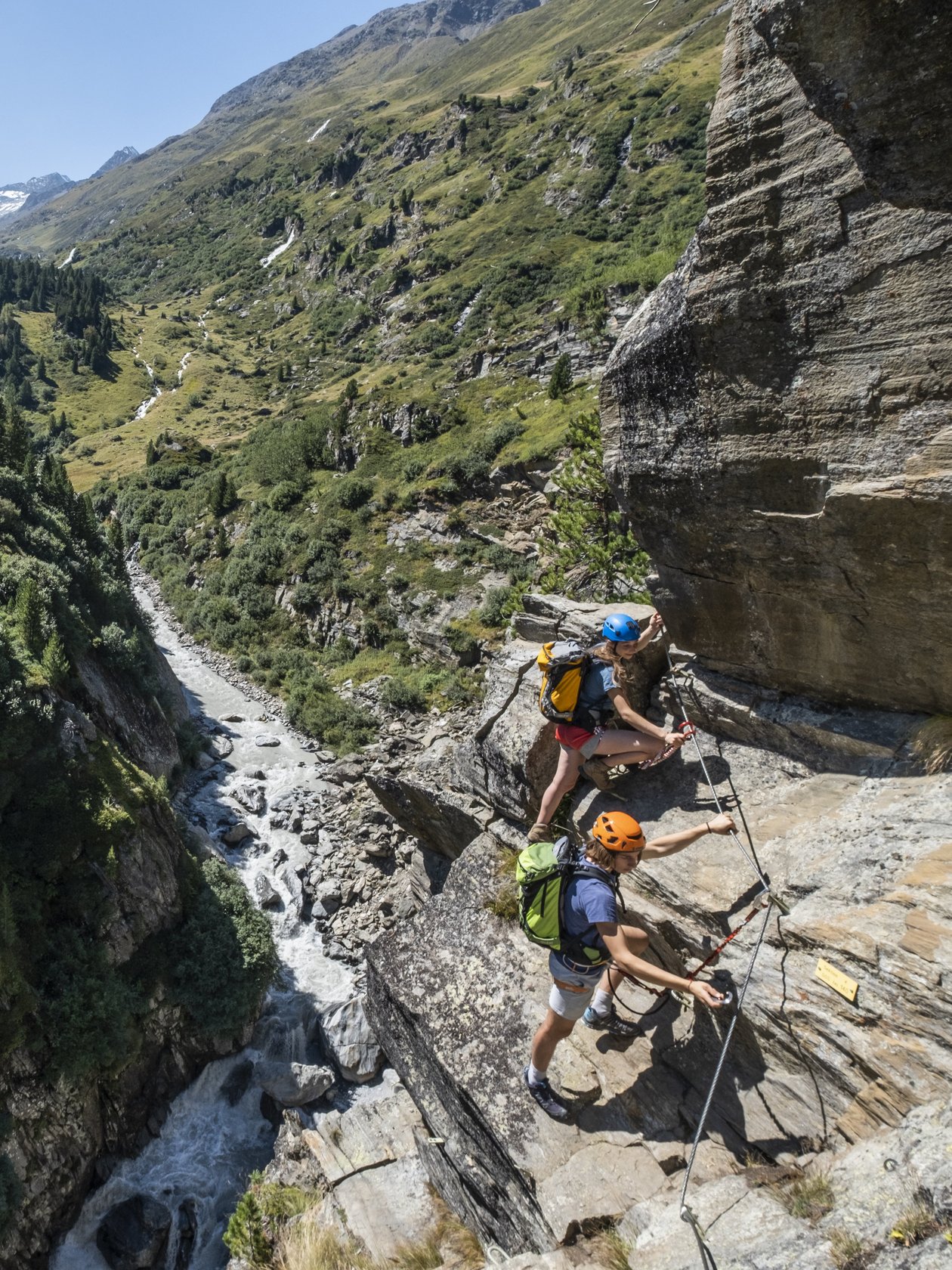 Two climbers navigate rocky terrain in a mountainous area with a river below and snow-capped peaks