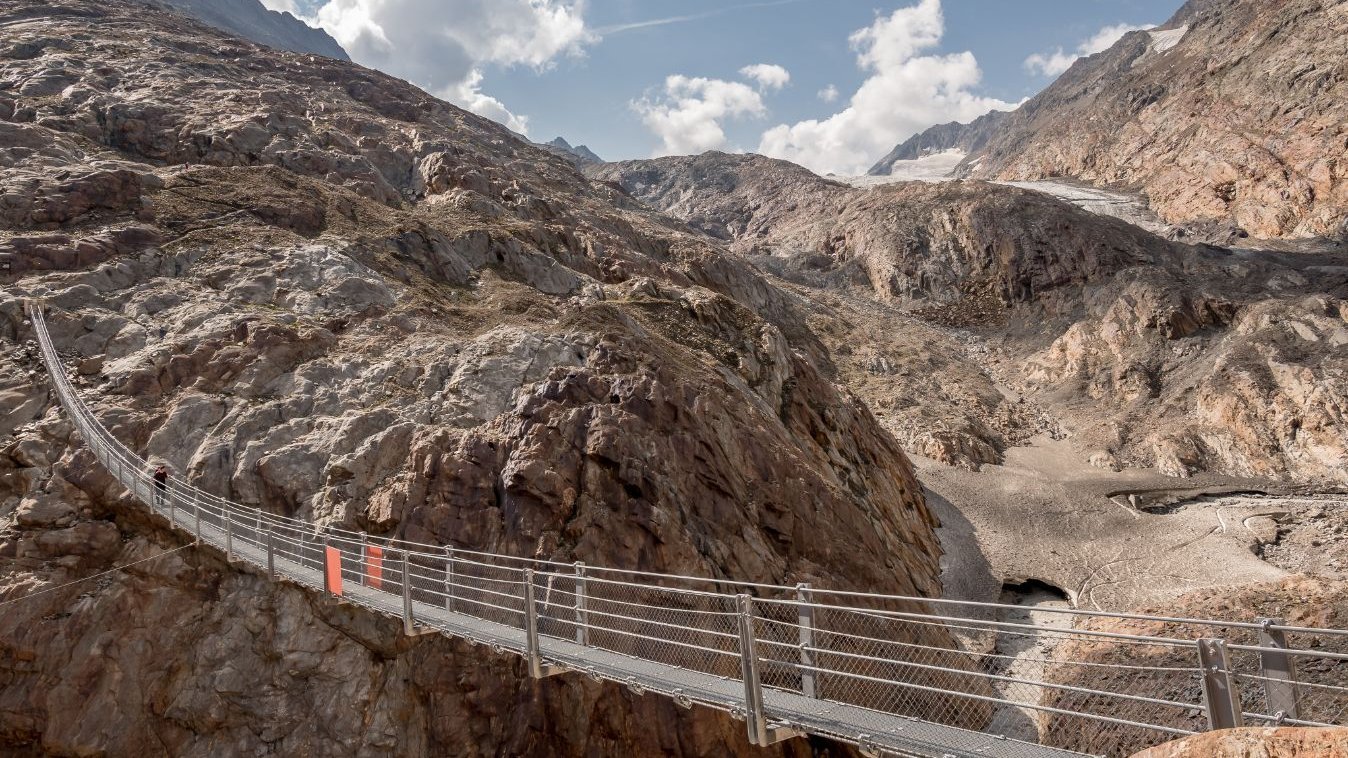 Schwebende Brücke in den Bergen Hängebrücke über felsige Landschaft mit Bergen und Gletscher im Hintergrund, ideal für Outdoor-Abenteuer.