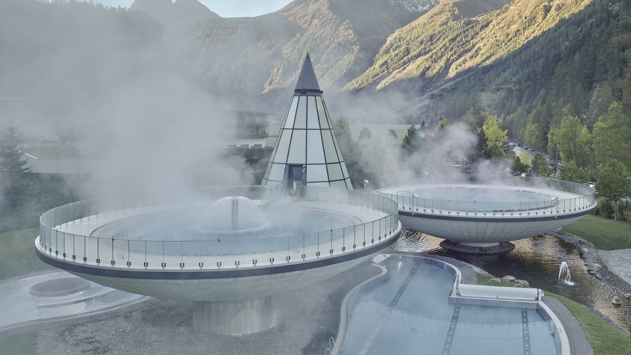 Moderner Spa-Bereich mit runden Gebäuden vor bergiger Landschaft und Nebel im Hintergrund.