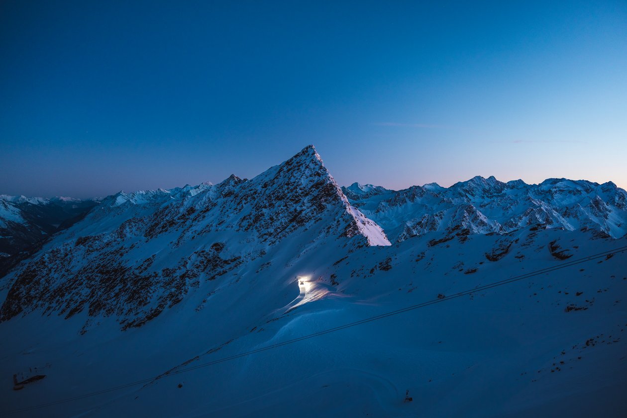 A snow-covered mountain peak glowing at sunset, surrounded by additional mountains in the background.