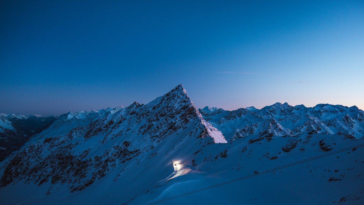 Schneeüberzogene Berge im Zwielicht, ein markanter Gipfel leuchtet künstlich auf.
