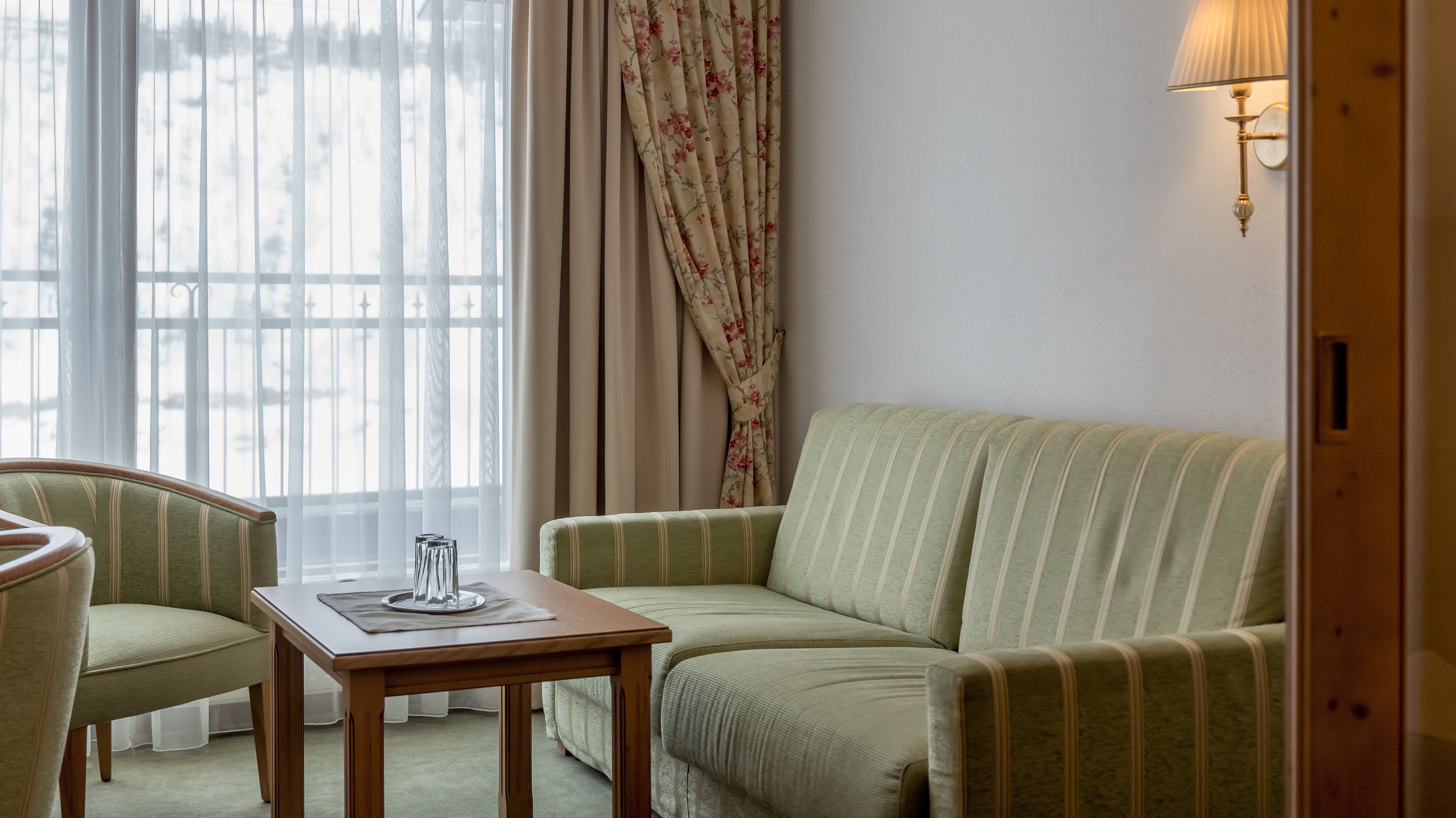 Hotelzimmer mit grün gestreiftem Sofa, Holztisch und großen Fenstern für Tageslicht.