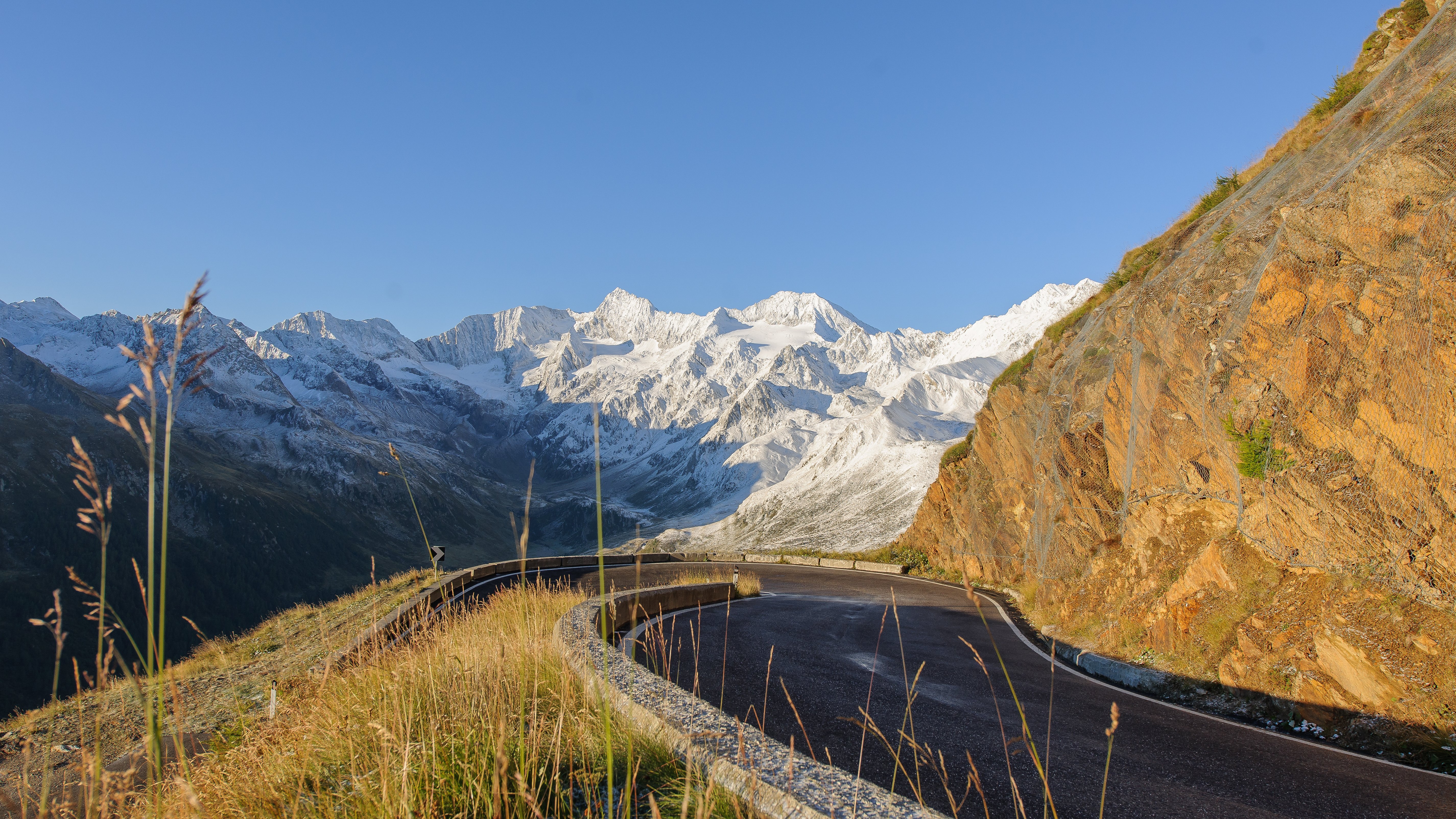 Eine kurvenreiche Straße, umgeben von schneebedeckten Gipfeln und grüner Natur unter einem klaren Himmel.