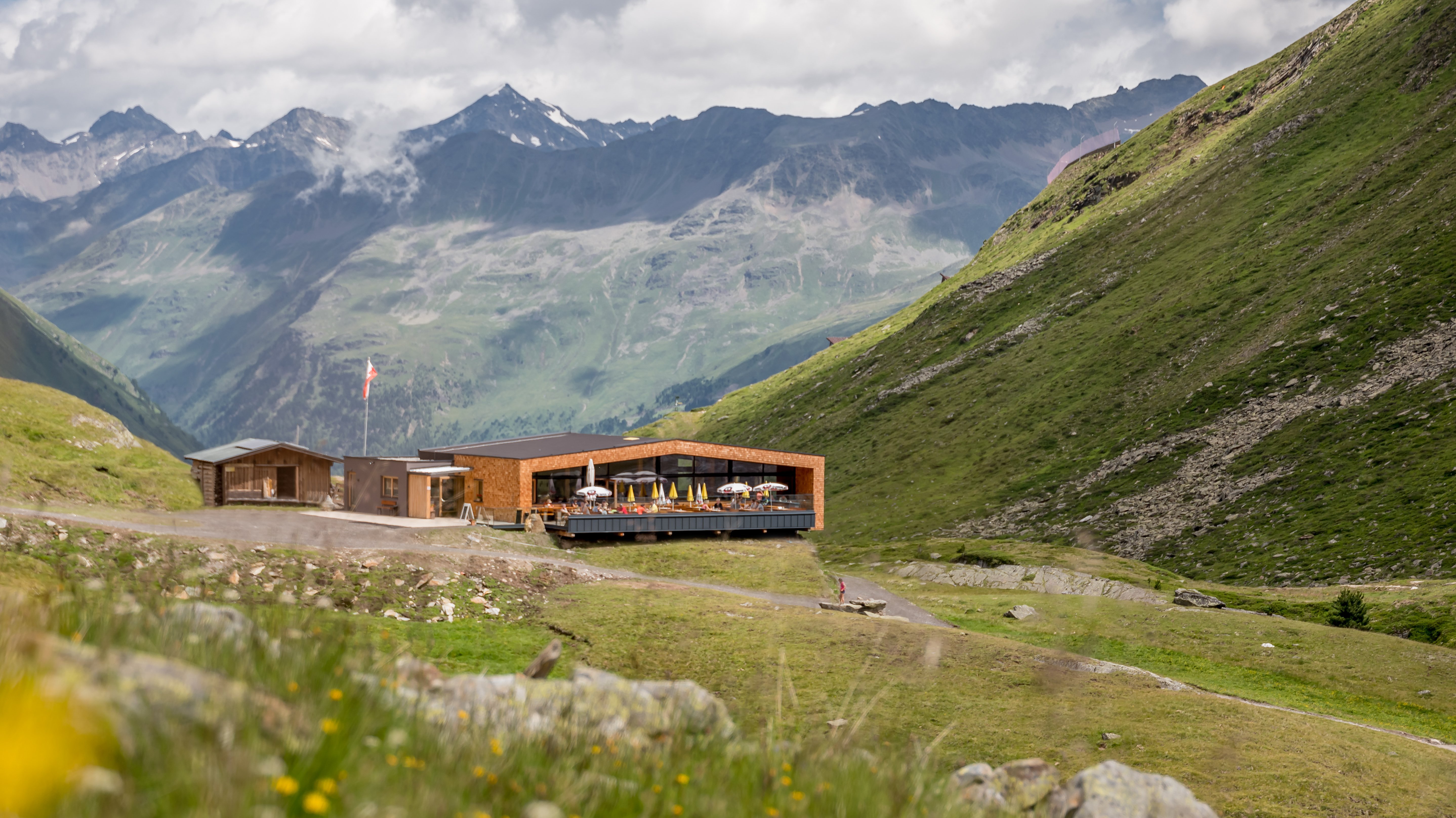 Moderne Berghütte im Grünen Blick auf eine moderne Berghütte in einem grünen Tal, umgeben von Bergen unter einem teils bewölkten Himmel.