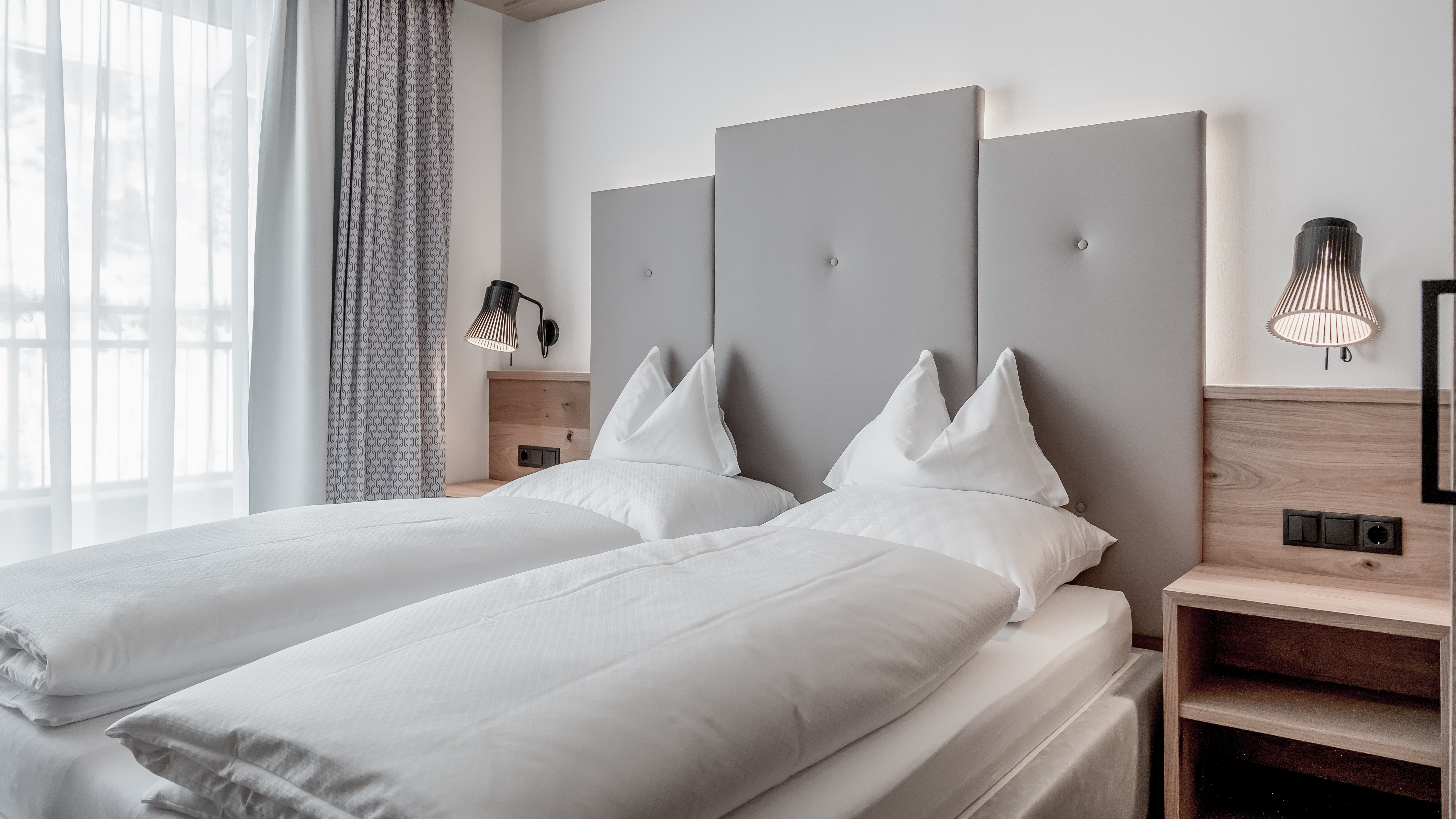 Moderne Unterkunft im Alpenland Zwei Einzelbetten mit weißer Bettwäsche in einem modernen Hotelzimmer mit Holz-Nachttisch und großen Fenstern.