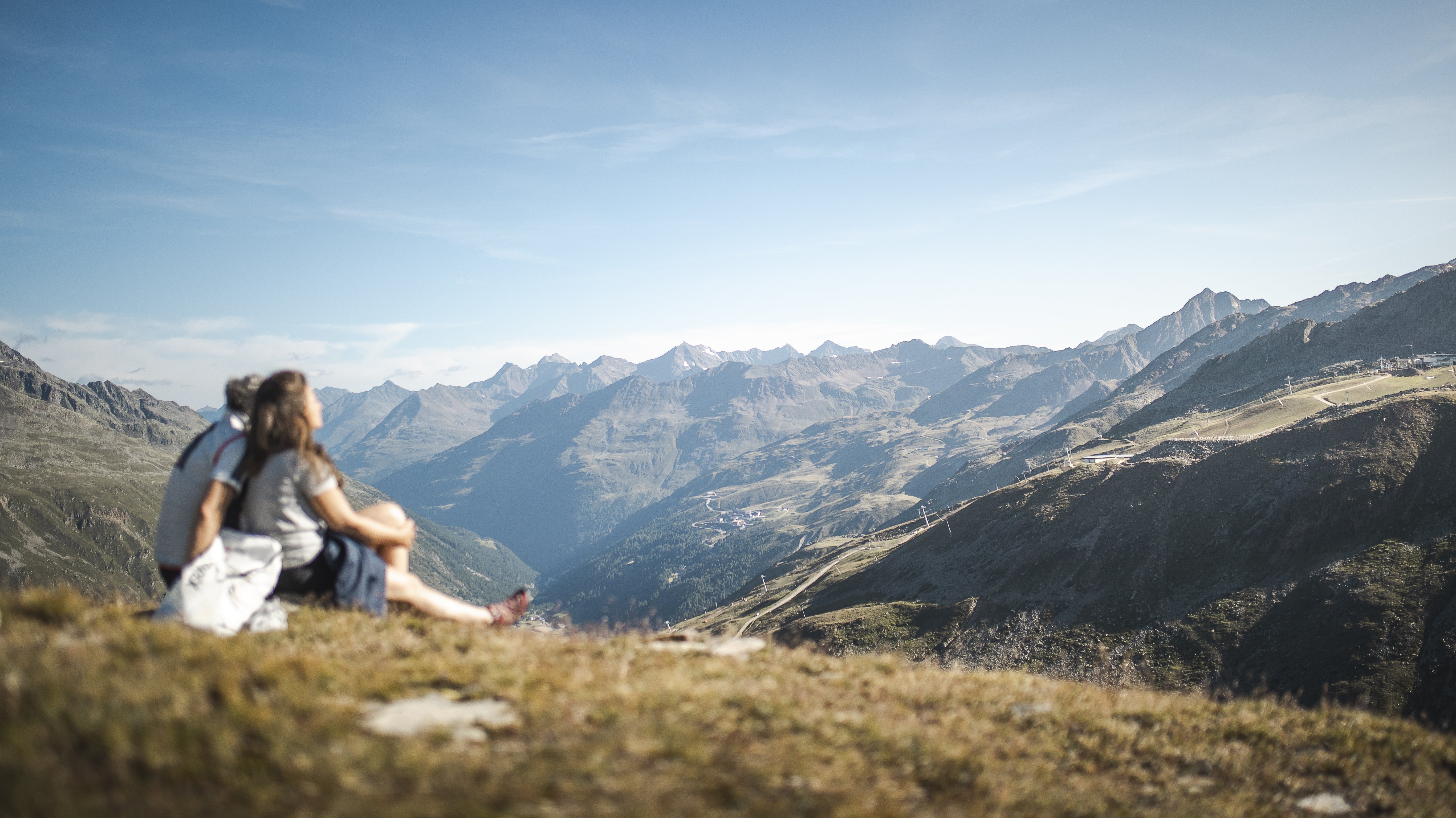 Ein Paar sitzt auf einer Wiese und genießt die Aussicht auf die Berglandschaft unter blauem Himmel.