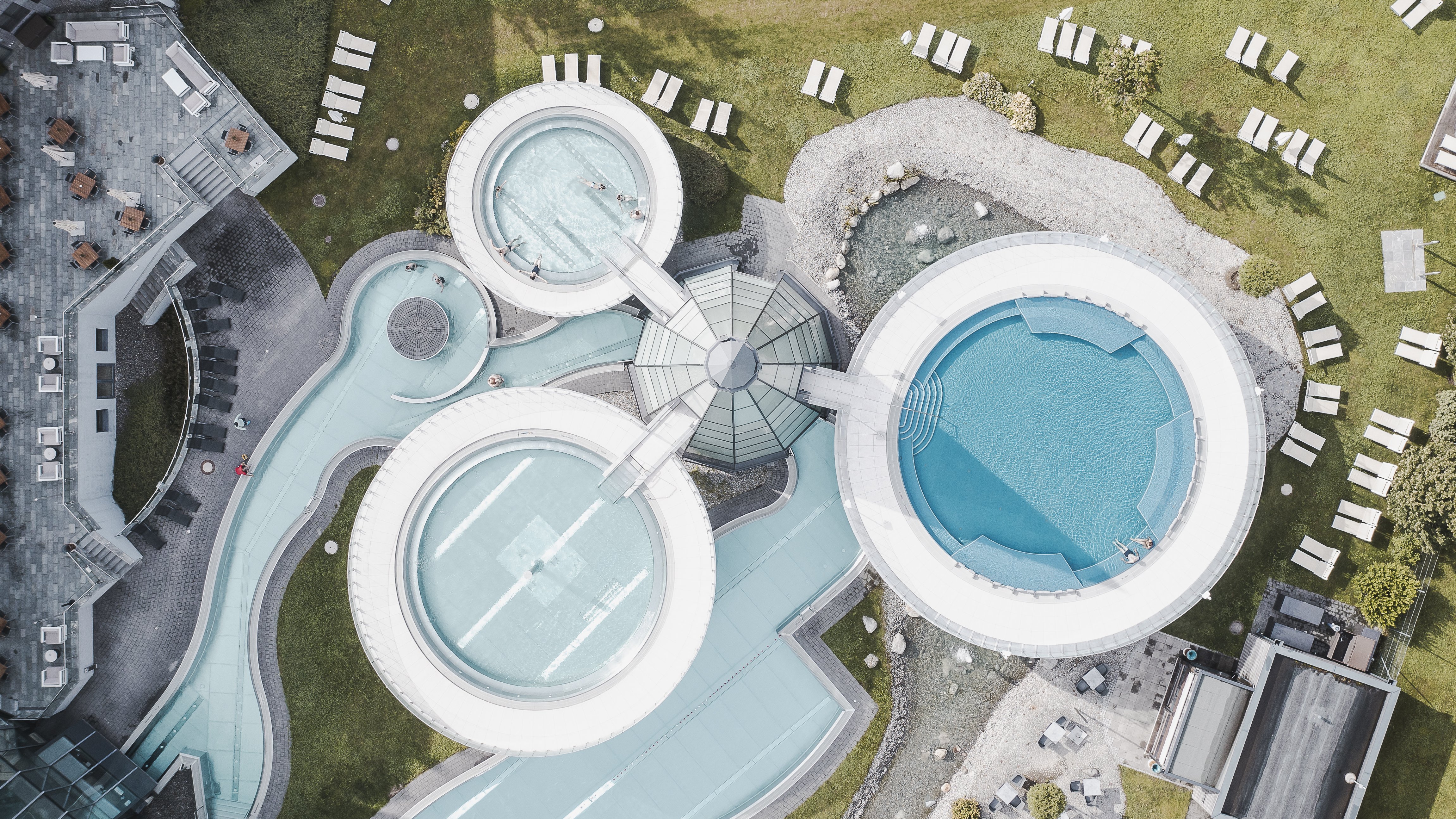 Moderne Schwimmbadanlage im Resort Luftaufnahme eines Resorts mit runden Schwimmbecken und Liegen auf grünem Rasen.