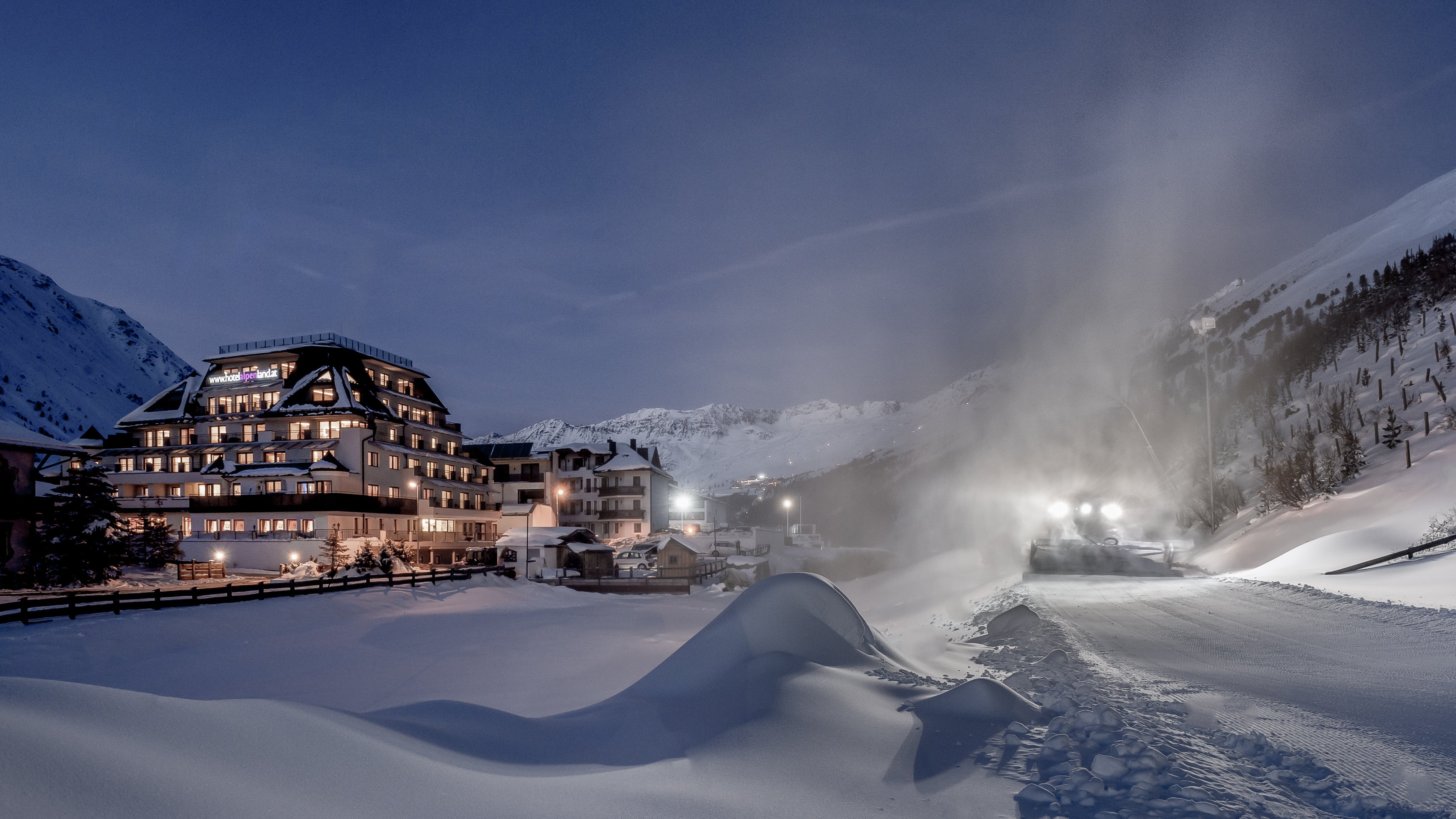 Winteridylle im Alpenland Hotel Alpenland in einem verschneiten Bergdorf bei Nacht, Schneepflug räumt die Straße.