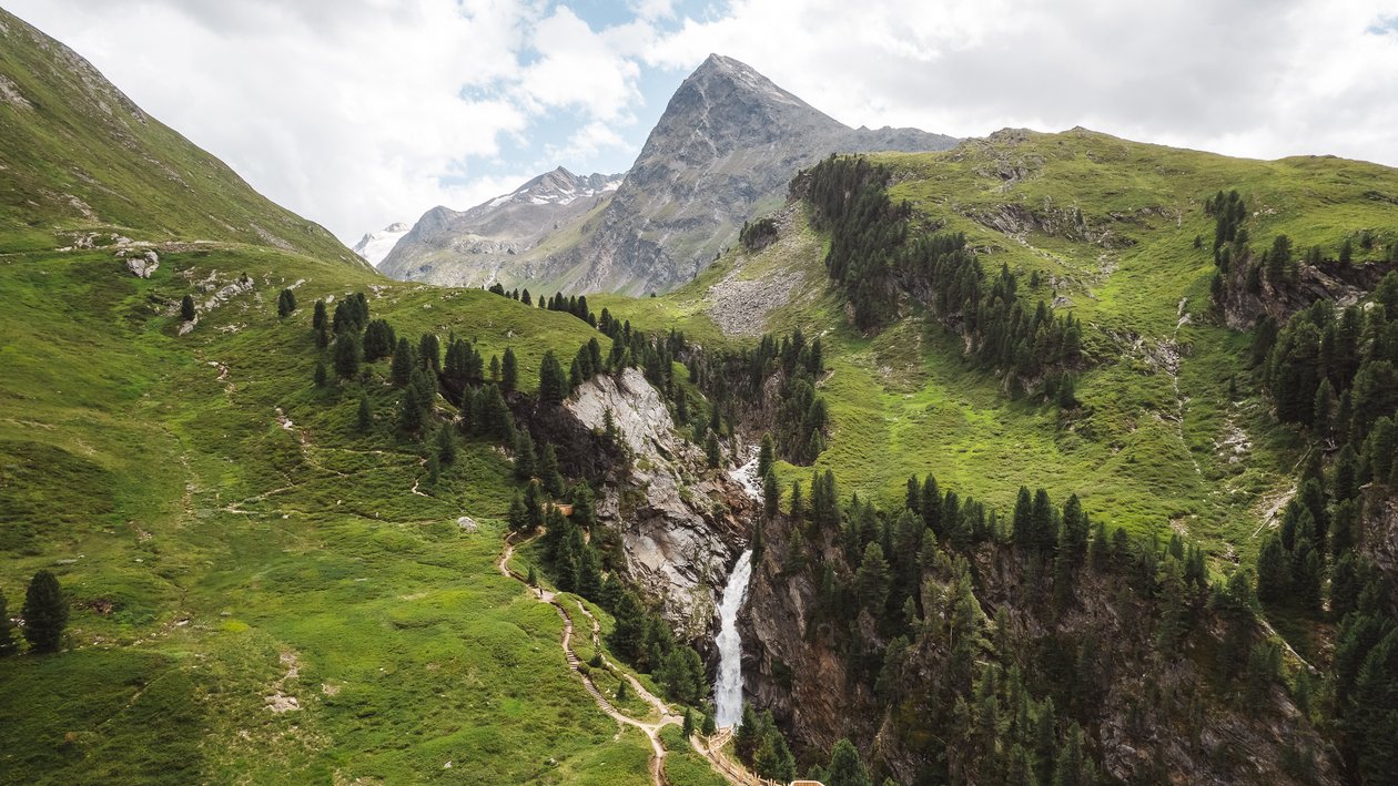 Ein Wasserfall fließt über einen steilen Felsen in einer grünen Berglandschaft mit Wanderwegen.