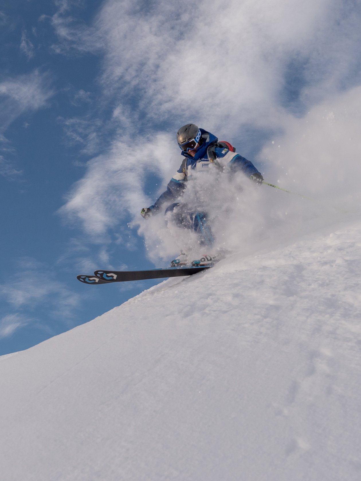 Ein Skifahrer fährt eine verschneite Piste hinunter und wirbelt Schnee auf, unter blauem Himmel.