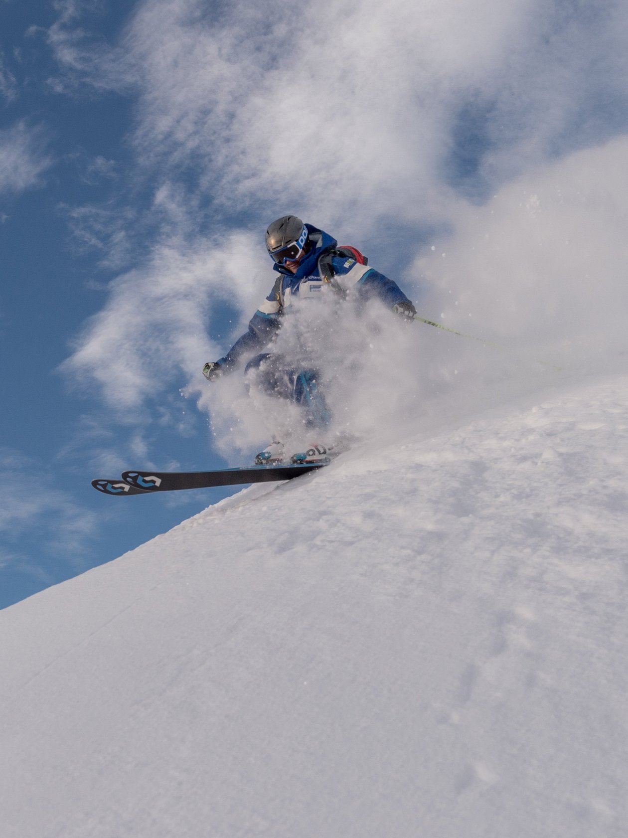 Ein Skifahrer fährt eine verschneite Piste hinunter und wirbelt Schnee auf, unter blauem Himmel.