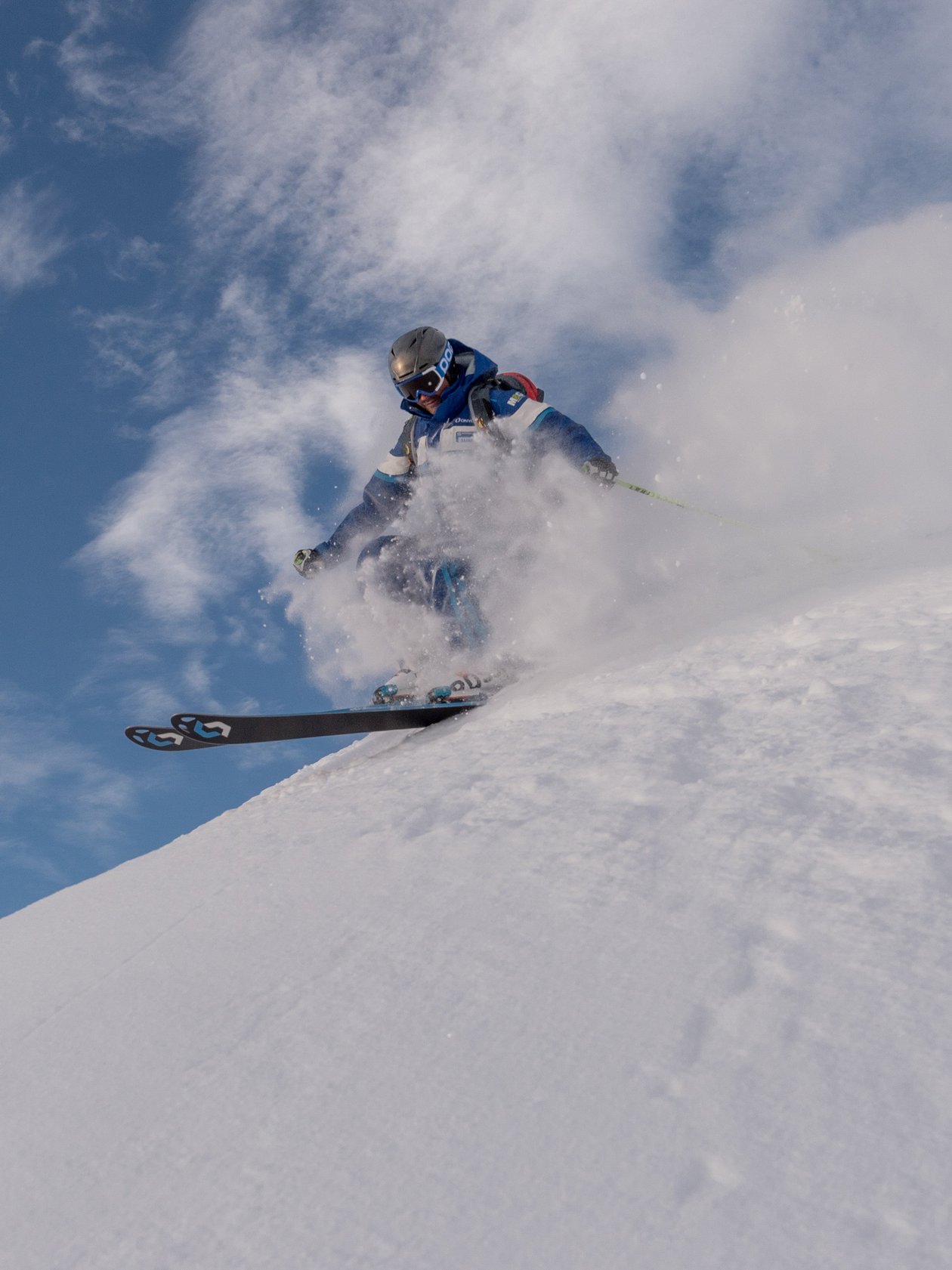 A skier races down a snowy slope, kicking up snow against a blue sky backdrop during winter sports activity.
