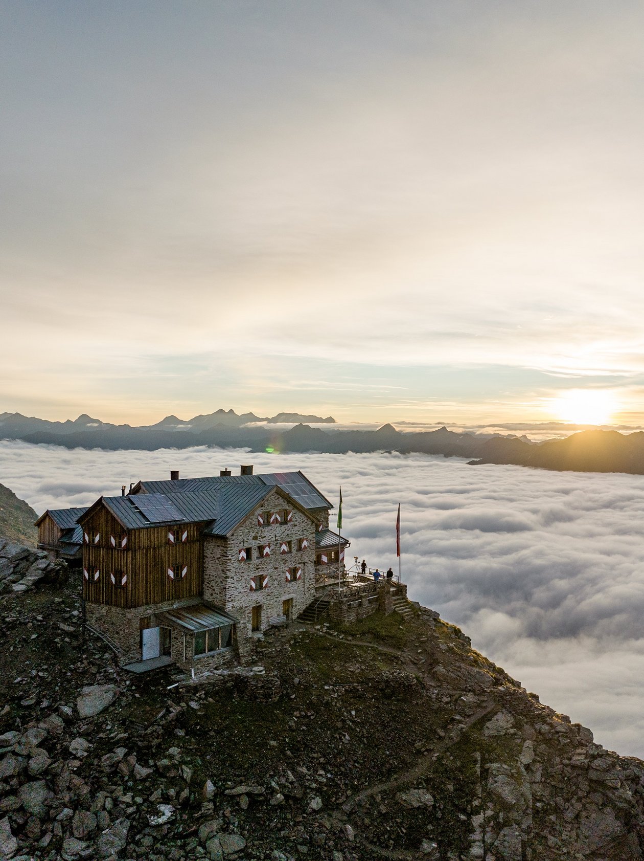 Mountain lodge on rocky terrain above clouds with a sunrise in the background and alpine scenery