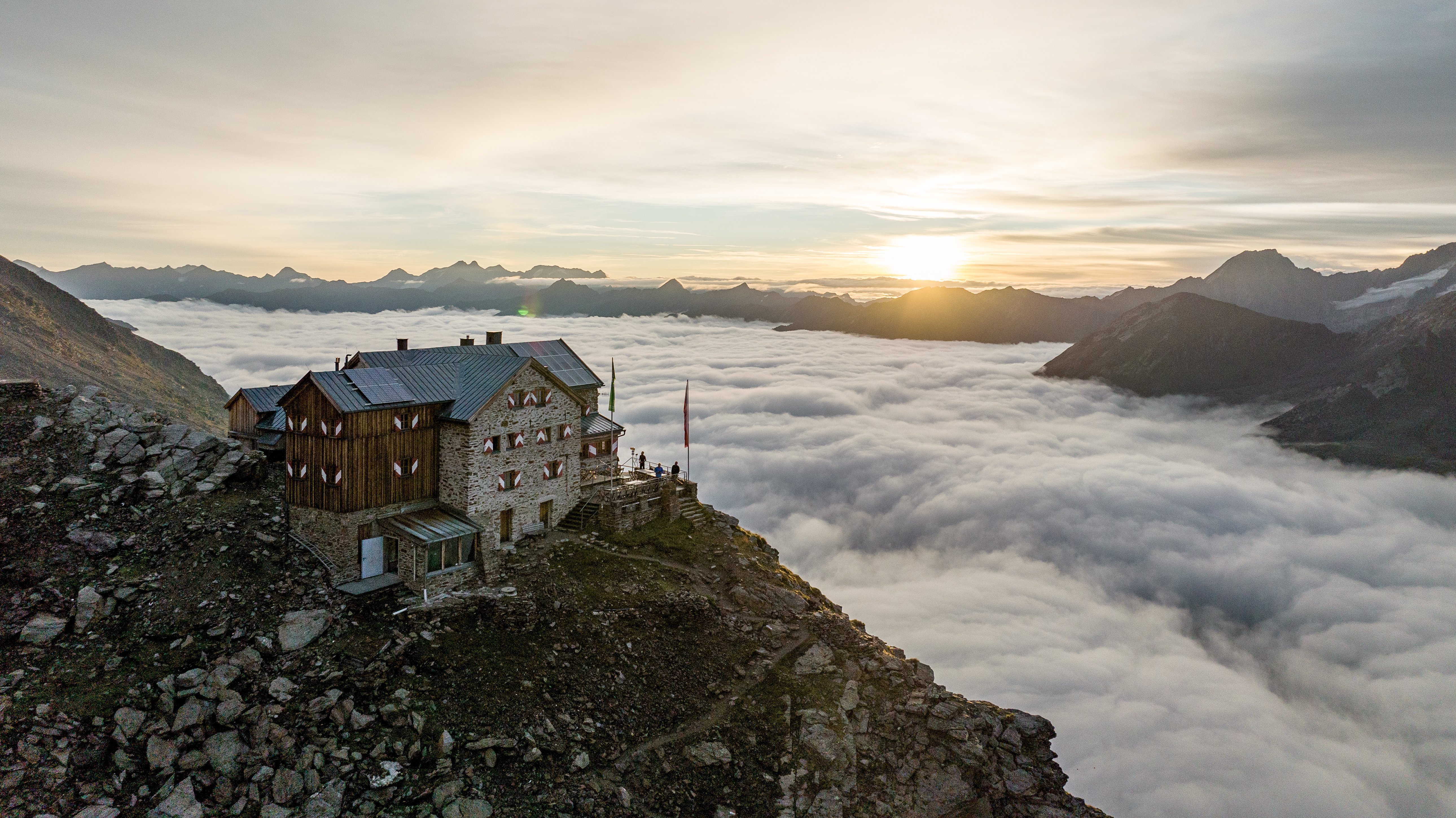 Berghütte auf steinigem Terrain mit Wolkenmeer und Sonnenuntergangshimmel im Hintergrund