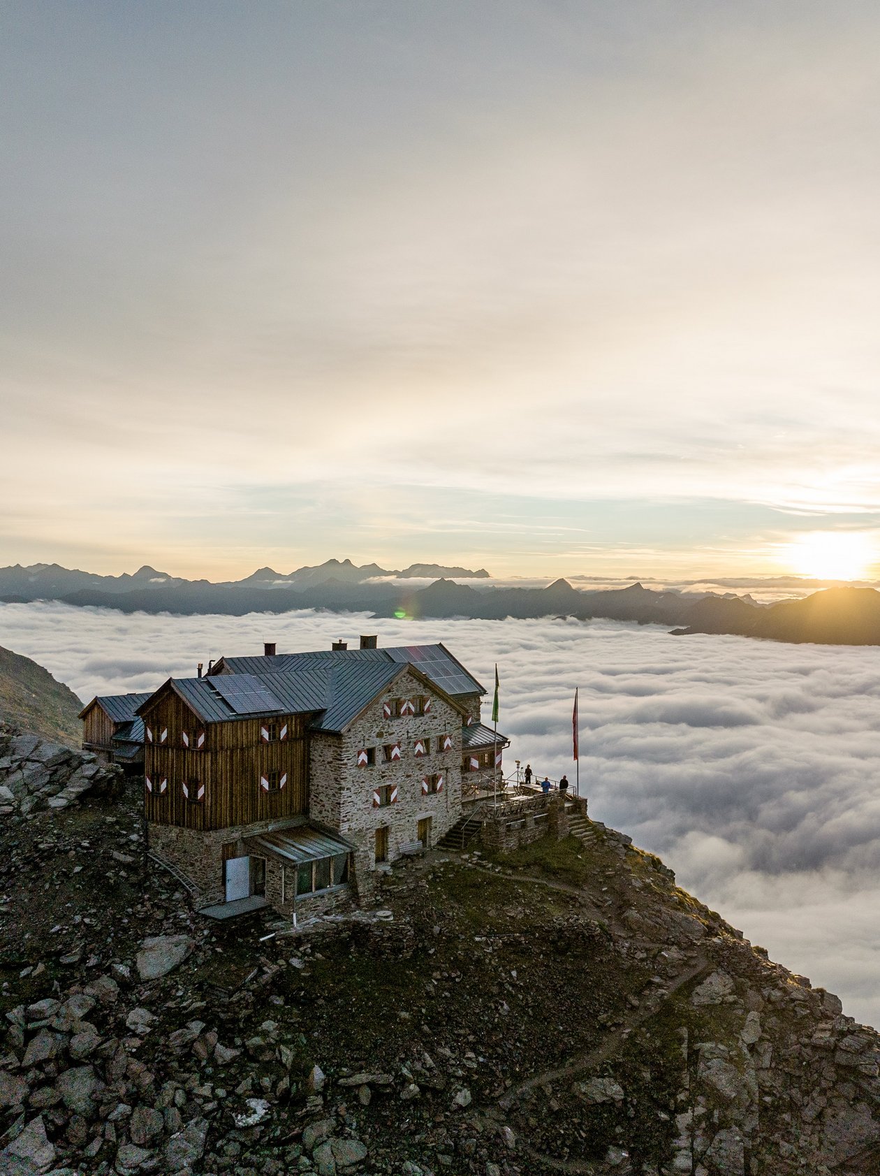 Alpenidyll im Sonnenuntergang Berghütte auf steinigem Terrain mit Wolkenmeer und Sonnenuntergangshimmel im Hintergrund