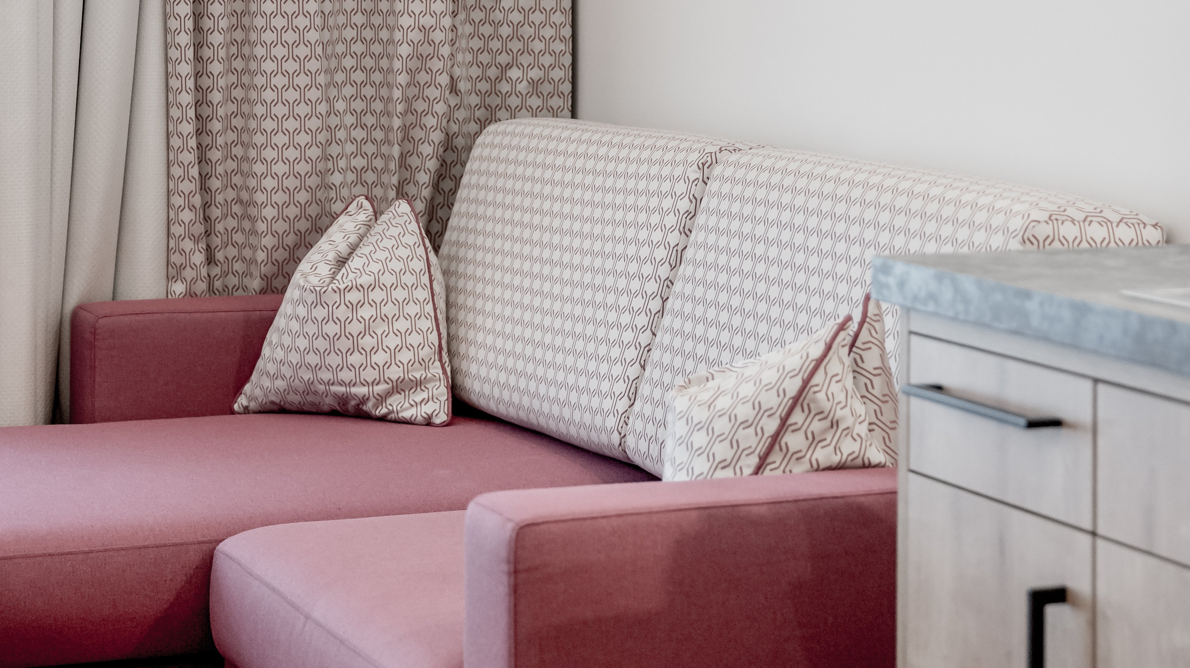 Gemütliches Wohnambiente im Alpenland Wohnzimmer-Ecke mit einem rosa Sofa und gemusterten Vorhängen sowie eingerahmten Fotografien an der Wand.