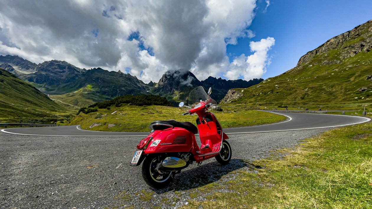 Eine rote Vespa steht auf einer kurvenreichen Bergstraße, umgeben von grünen Hügeln und bewölktem Himmel.