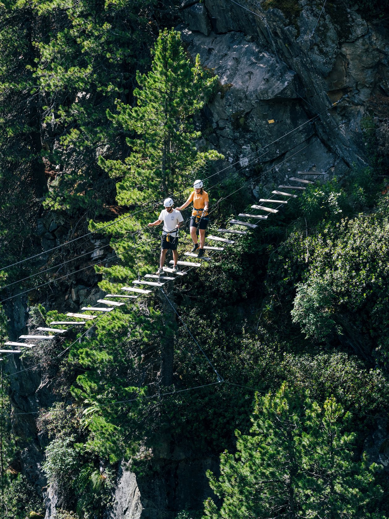 Zwei Wanderer gehen über eine Hängebrücke, umgeben von Bäumen und Bergen in der Natur.