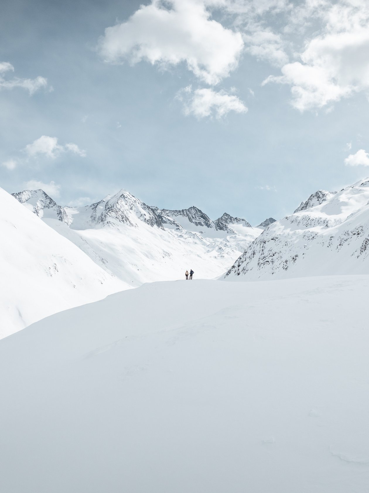 Schneelandschaft mit Bergkulisse Zwei Personen stehen im Schnee vor hohen Bergen unter einem teils bewölkten Himmel.