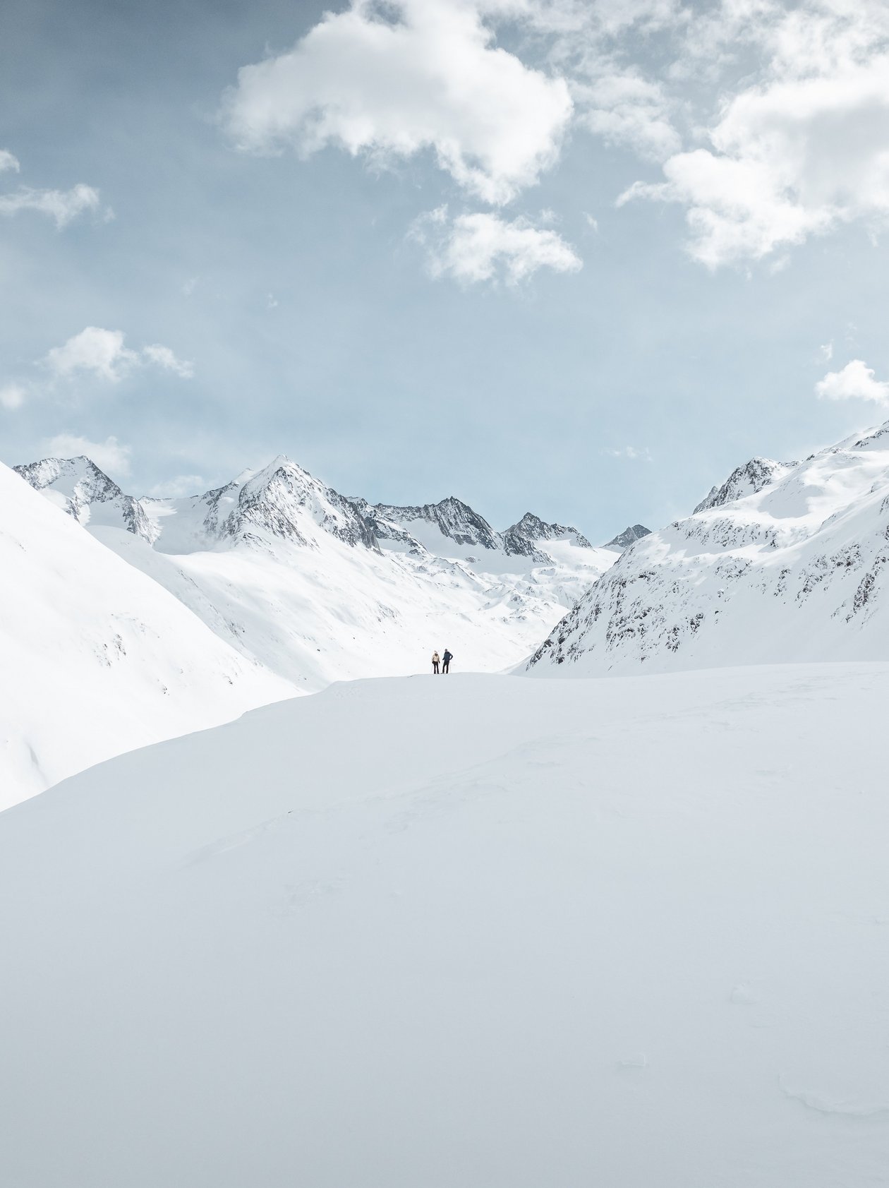 Embrace the Alpine Adventure Two people standing on snowy ground surrounded by high mountains under a partly cloudy sky