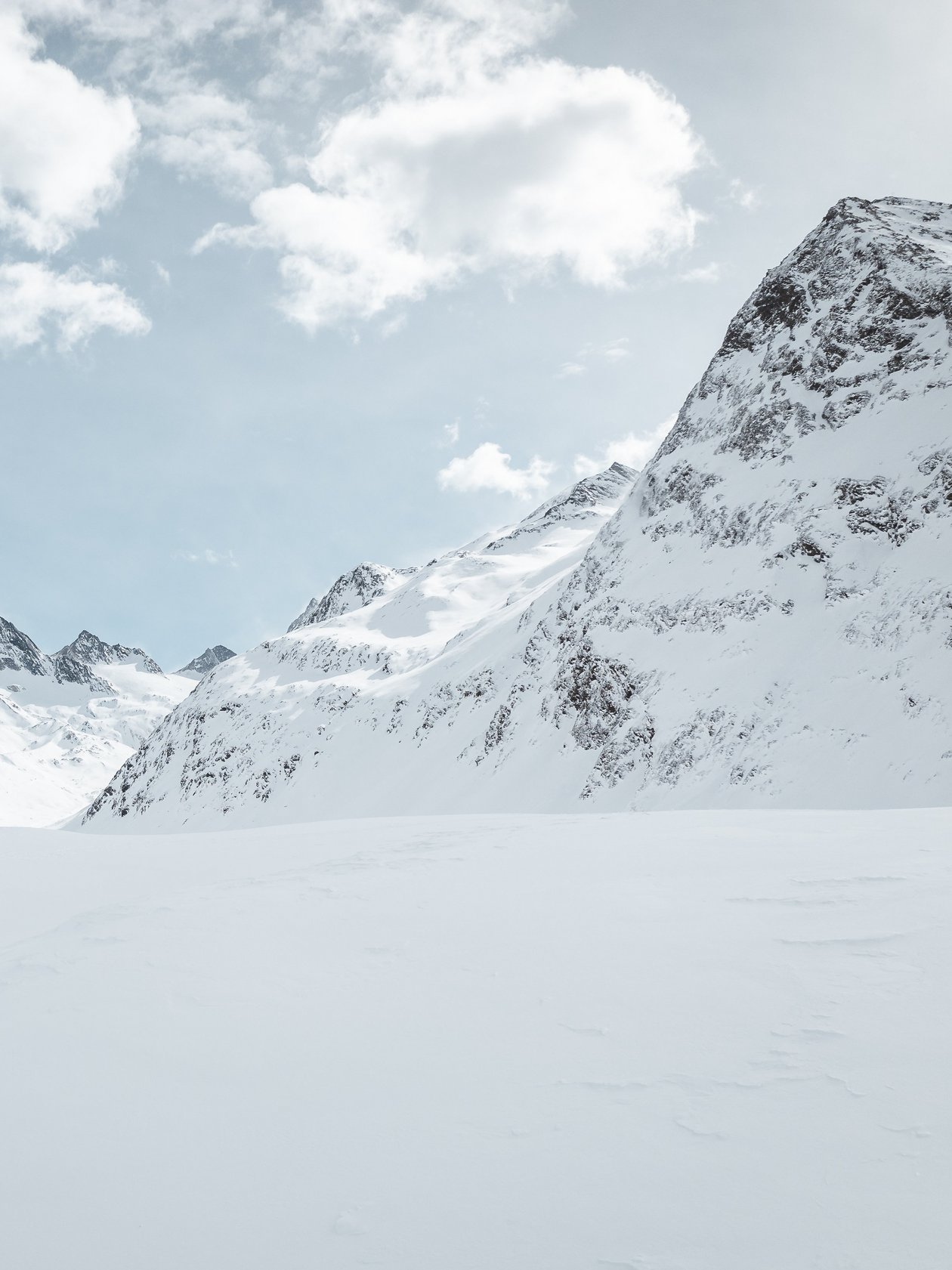 Zwei Personen stehen im Schnee vor hohen Bergen unter einem teils bewölkten Himmel.