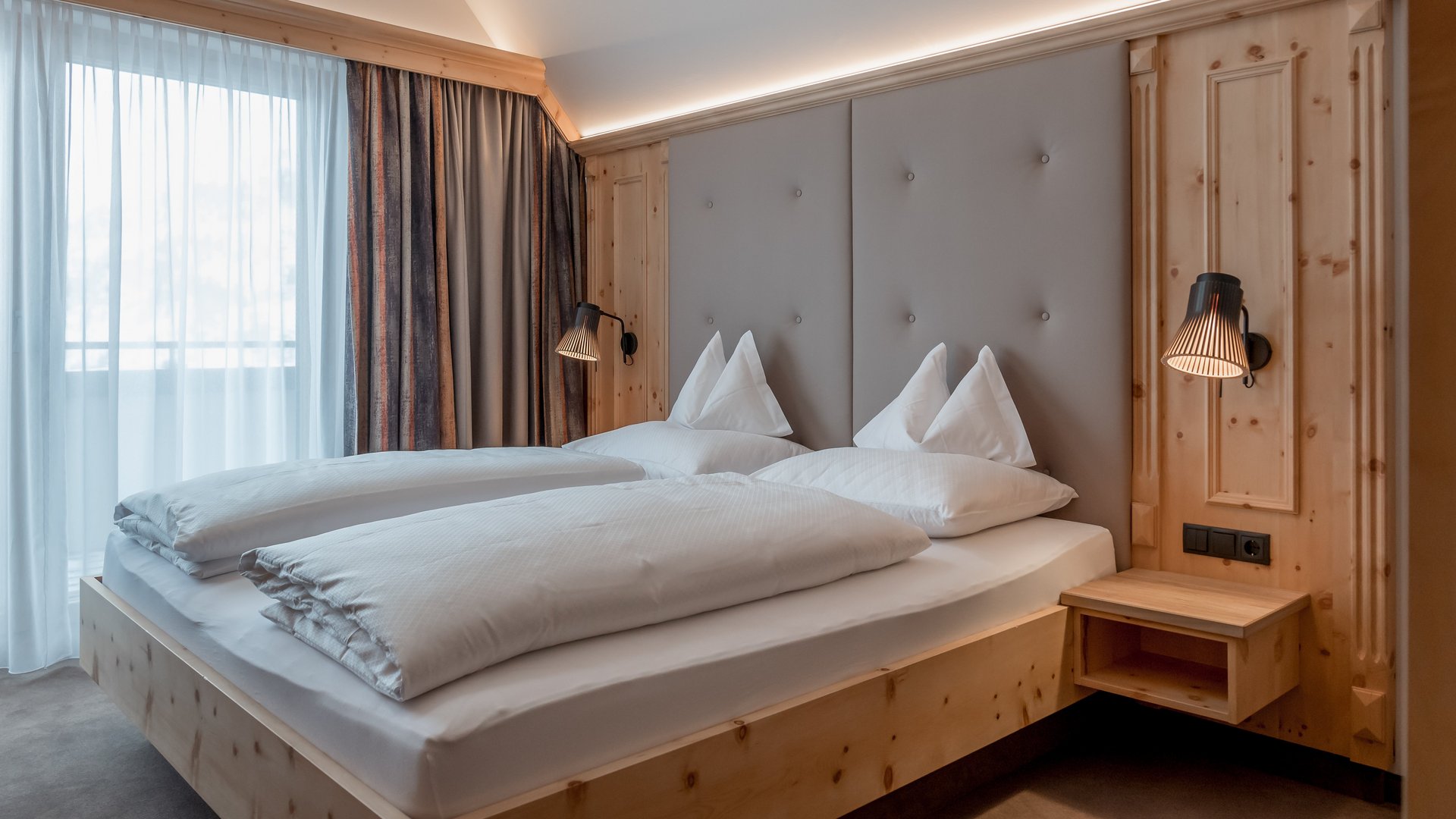 Modernes Schlafzimmer mit Doppelbett und Holzmöbeln in einem Hotelzimmer, große Fenster mit lichtdurchlässigen Vorhängen.