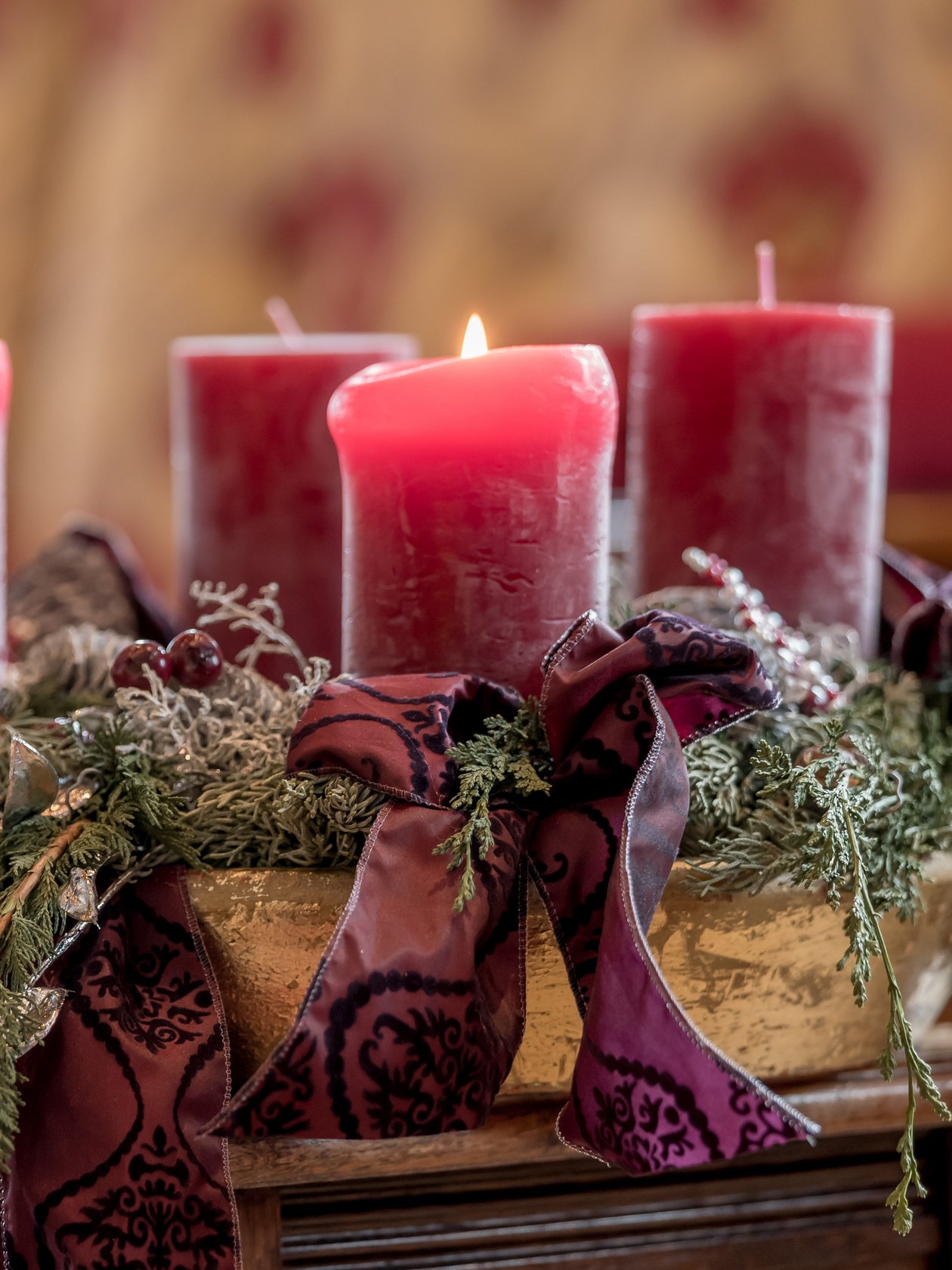 Vier rote Kerzen auf einem Adventskranz mit grünem Laub und Bändern in einem gemütlichen Raum.
