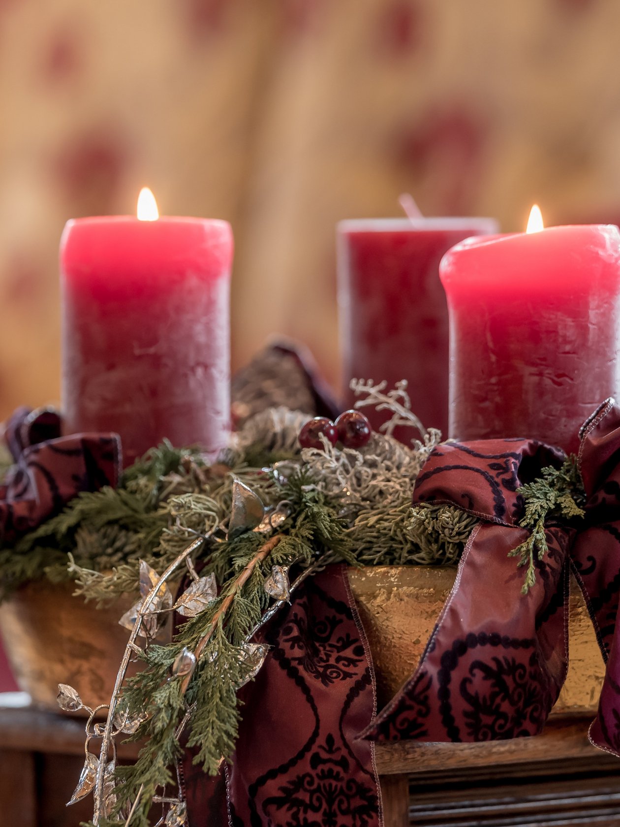 Vier rote Kerzen auf einem Adventskranz mit grünem Laub und Bändern in einem gemütlichen Raum.