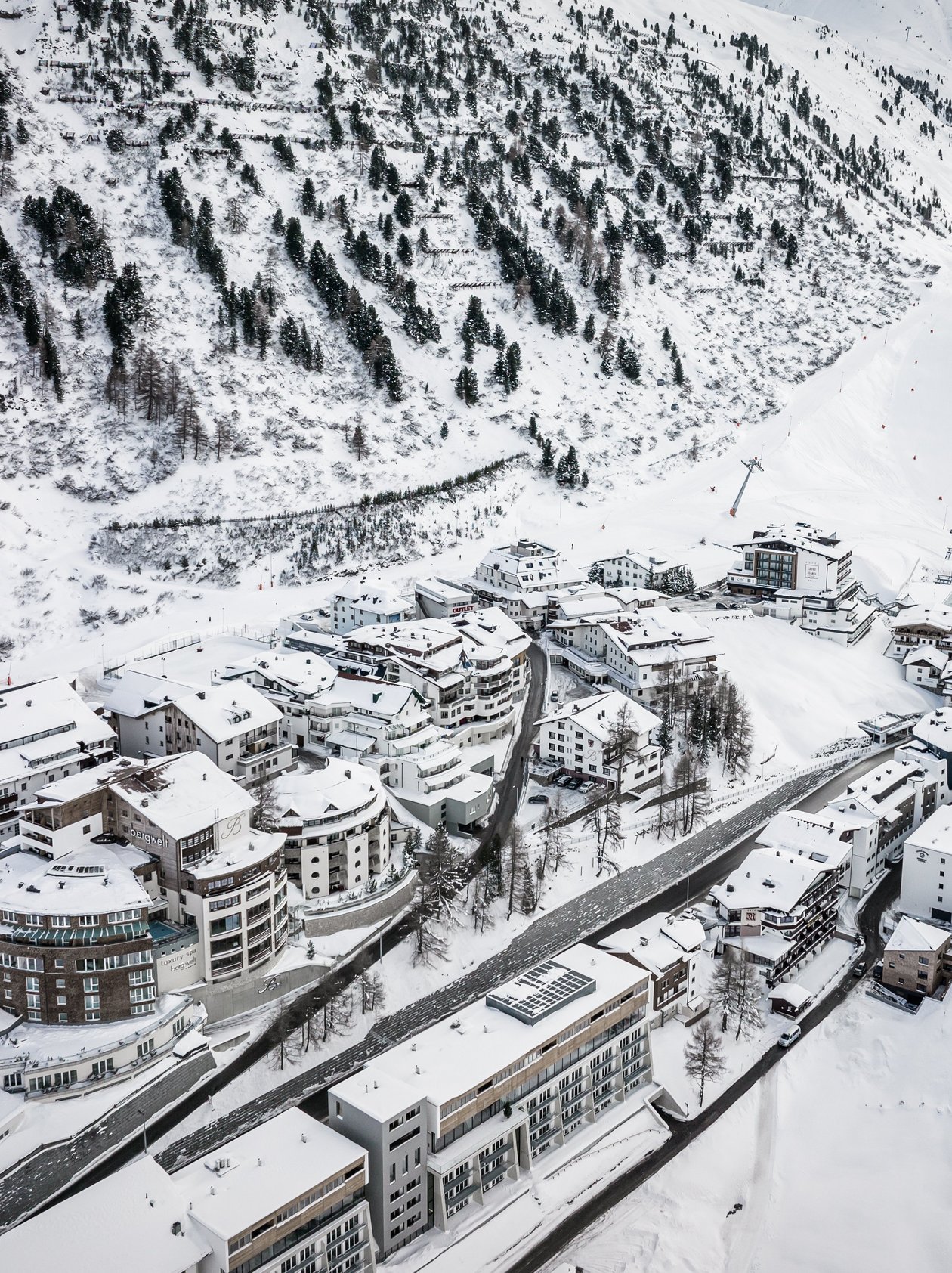 Luftaufnahme eines verschneiten Berghoteldorfs umgeben von Tannen und Pisten im Winter.