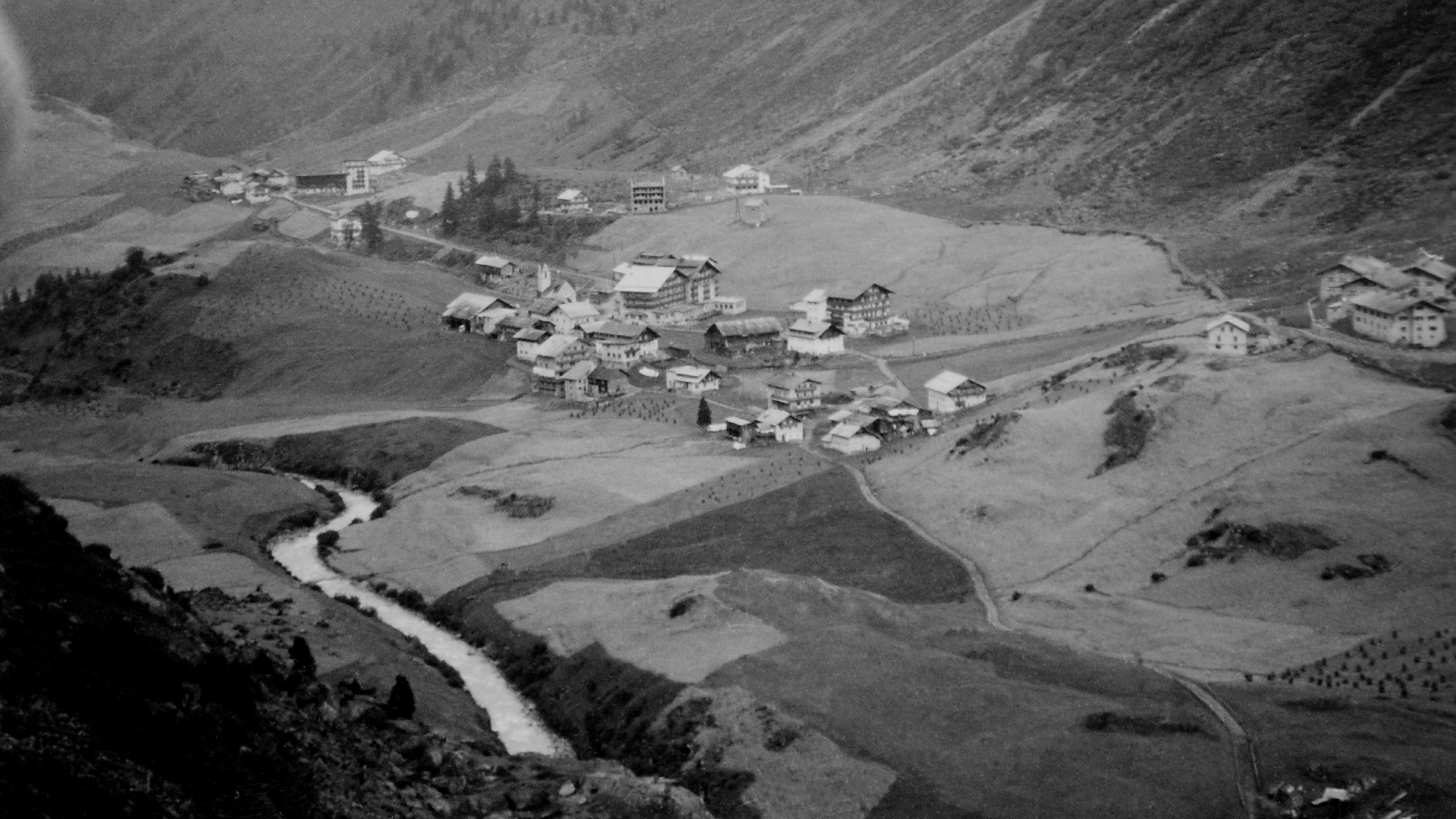 Bergdorf im Tal umgeben von Hügeln und einem Fluss in Schwarz-Weiß-Fotografie mit ländlichem Charakter.