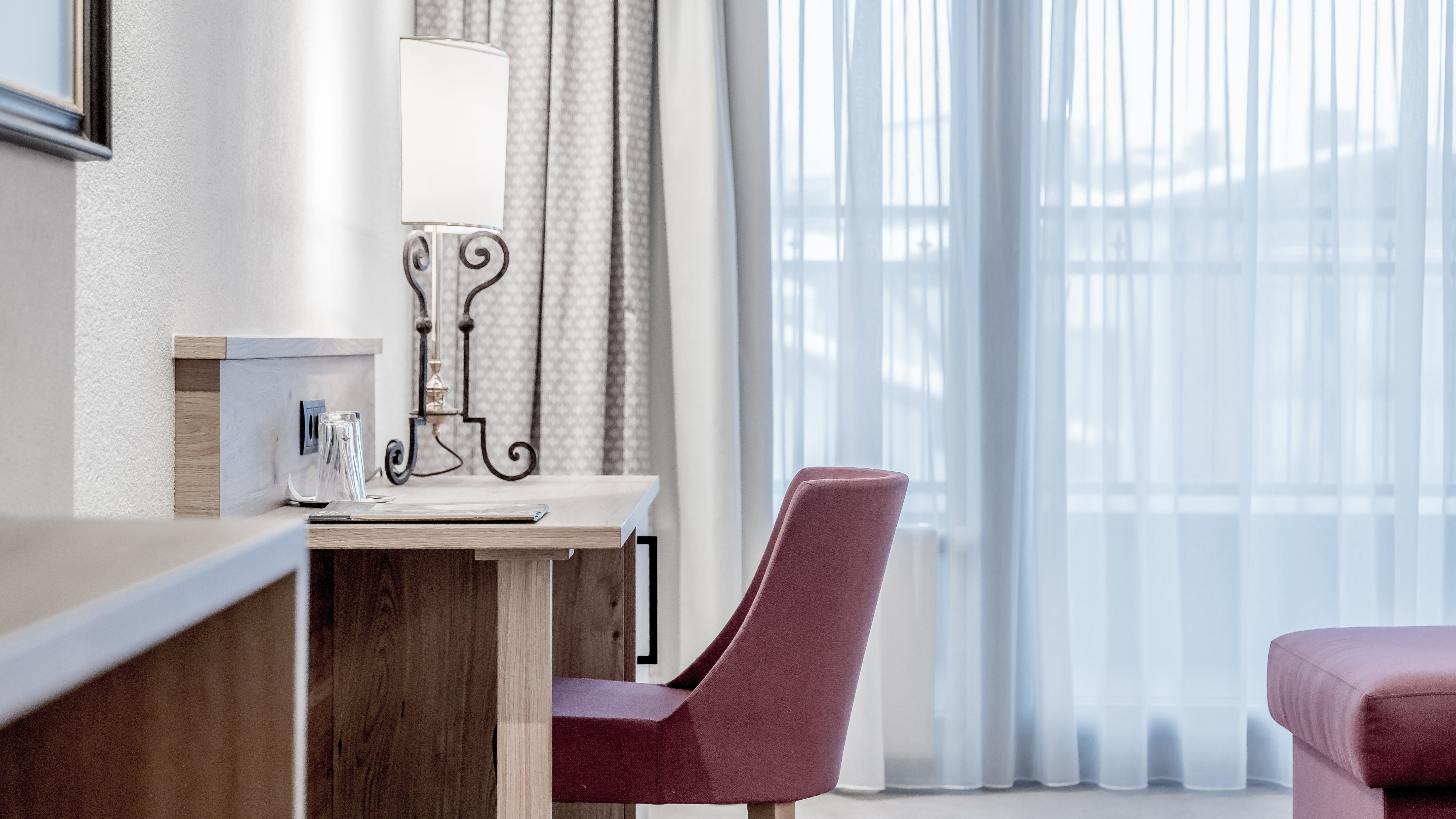 Moderne Eleganz in der Suite Helles Hotelzimmer mit Schreibtisch, Lampe und einem pinken Stuhl neben sheer Vorhängen.