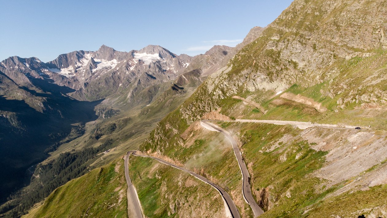 Luftaufnahme einer kurvigen Bergstraße umgeben von grünen Hügeln und Bergen im Hintergrund