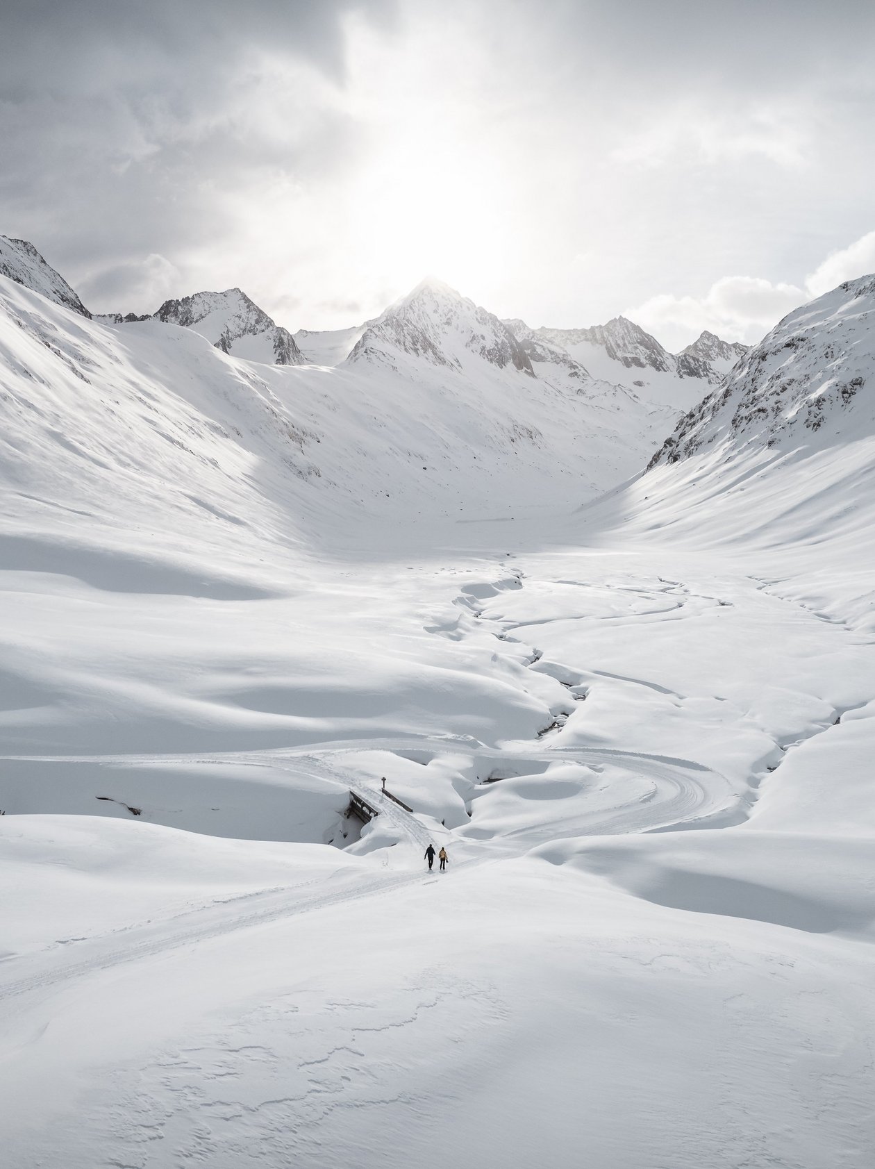 Serene snowy valley adventure Two figures hike through a snowy valley surrounded by mountains and a winding river of snow