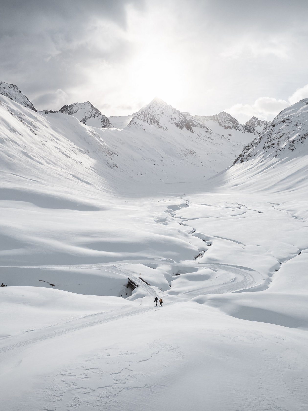Winterlandschaft im Ötztal Schneebedeckte Landschaft mit hohen Bergen und einem gewundenen Pfad, kleine Figuren sind in der Ferne zu sehen.
