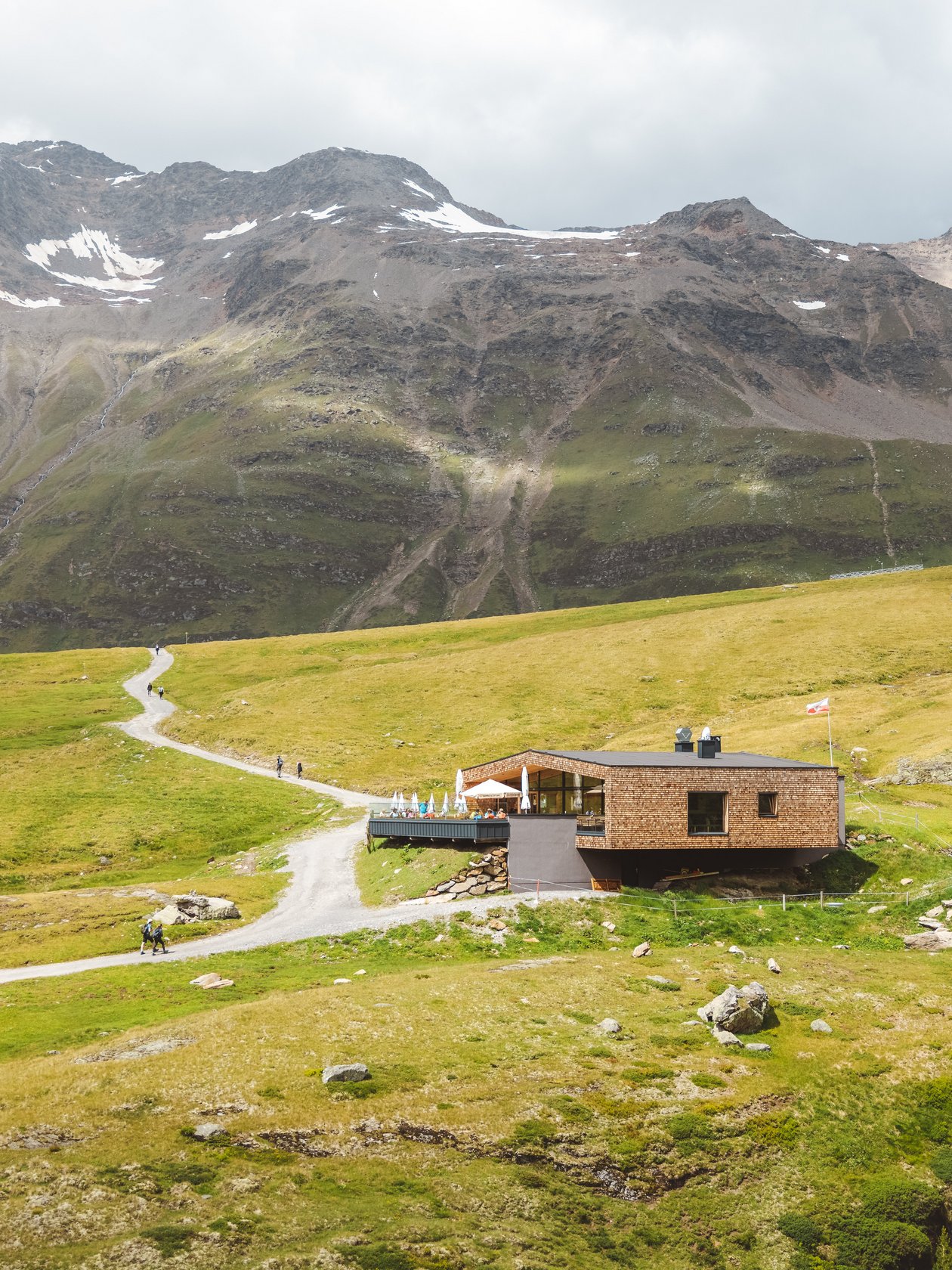 Holzhütte in einer bergigen Landschaft mit grünem Gras und felsigem Terrain unter einem bewölkten Himmel.